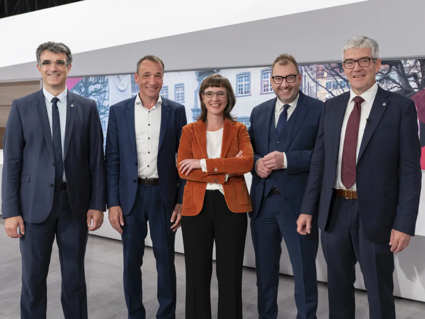 Cinque persone in abbigliamento formale sorridono insieme su uno sfondo moderno. Due uomini indossano completi blu, mentre una donna è in giacca arancione e pantaloni bianchi. Gli altri due uomini indossano completi scuri.