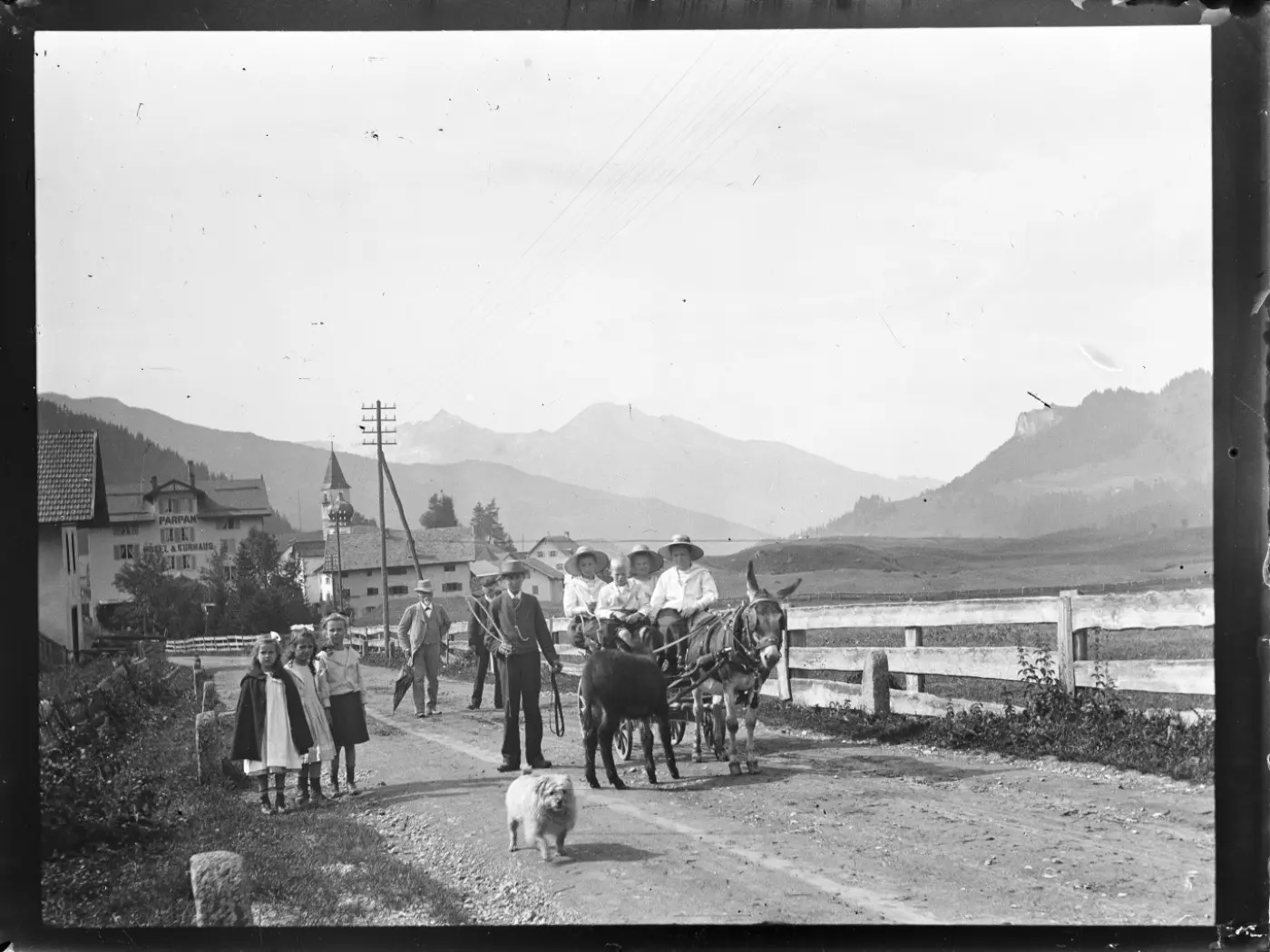 Due persone su un carro trainato da un asino, con due bambini e un cane accanto. Sullo sfondo, montagne e un paesaggio rurale. Una casa e alberi lungo la strada sterrata.