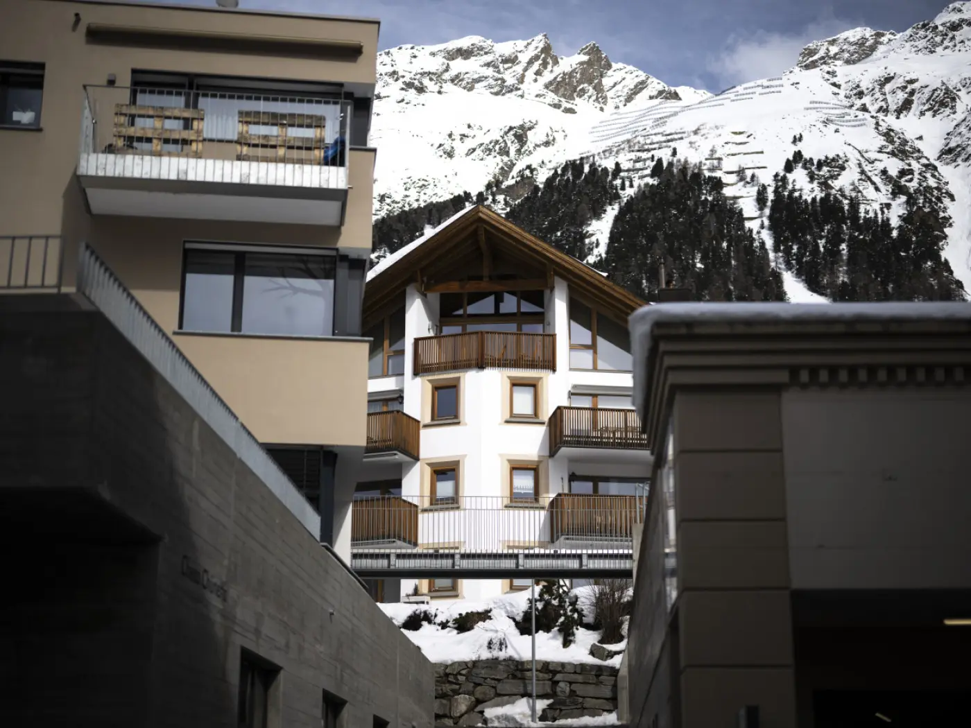 Una vista di un edificio architettonico con un tetto a spiovente, circondato da montagne innevate e un cielo parzialmente nuvoloso. Altri edifici moderni si trovano in primo piano.