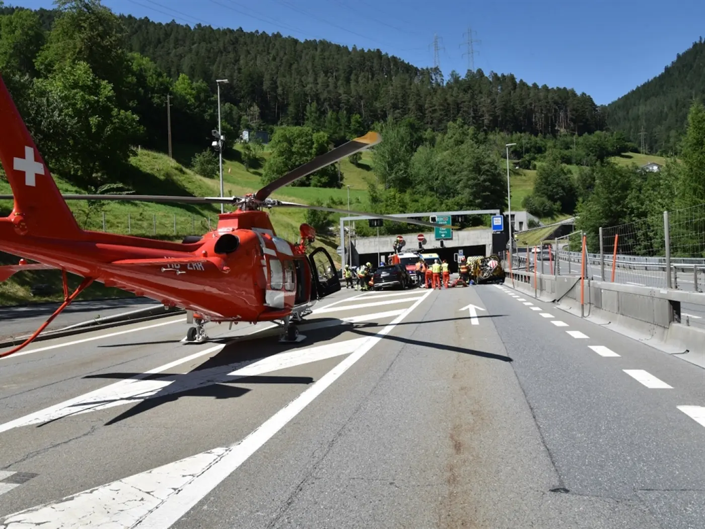Un elicottero rosso dei soccorsi atterrato su una strada vicino a un tunnel, con squadre di intervento che assistono in un'operazione di emergenza. Sullo sfondo, boschi verdi e segnaletica stradale visibile.