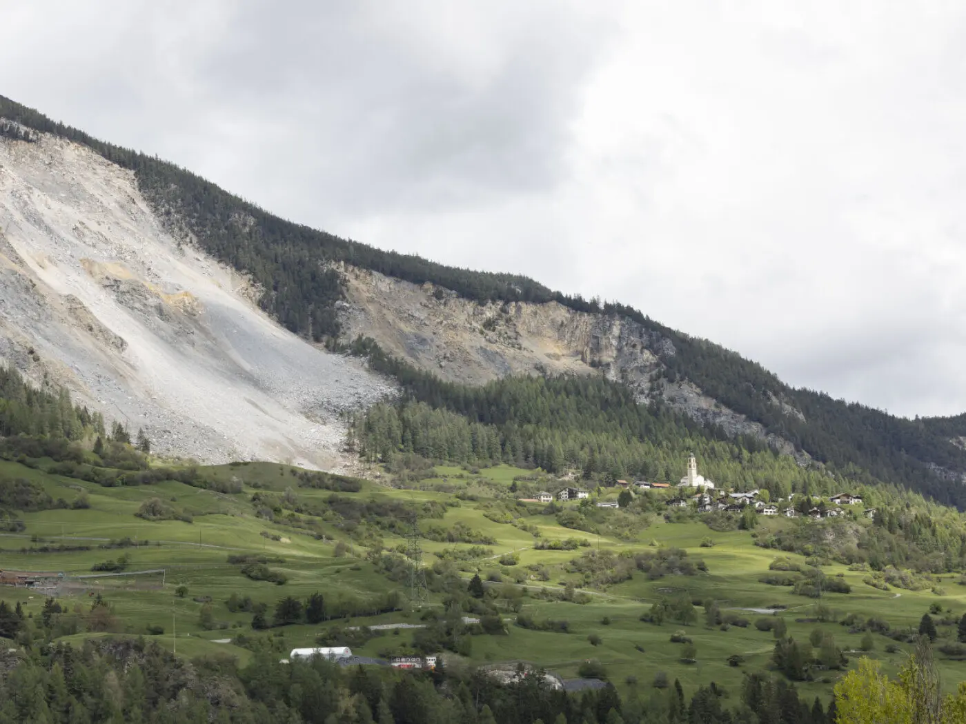 Un paesaggio montano con una collina verdeggiante, un piccolo villaggio visibile in basso e una grande parete rocciosa che mostra segni di frana. Nuvole grigie adornano il cielo.