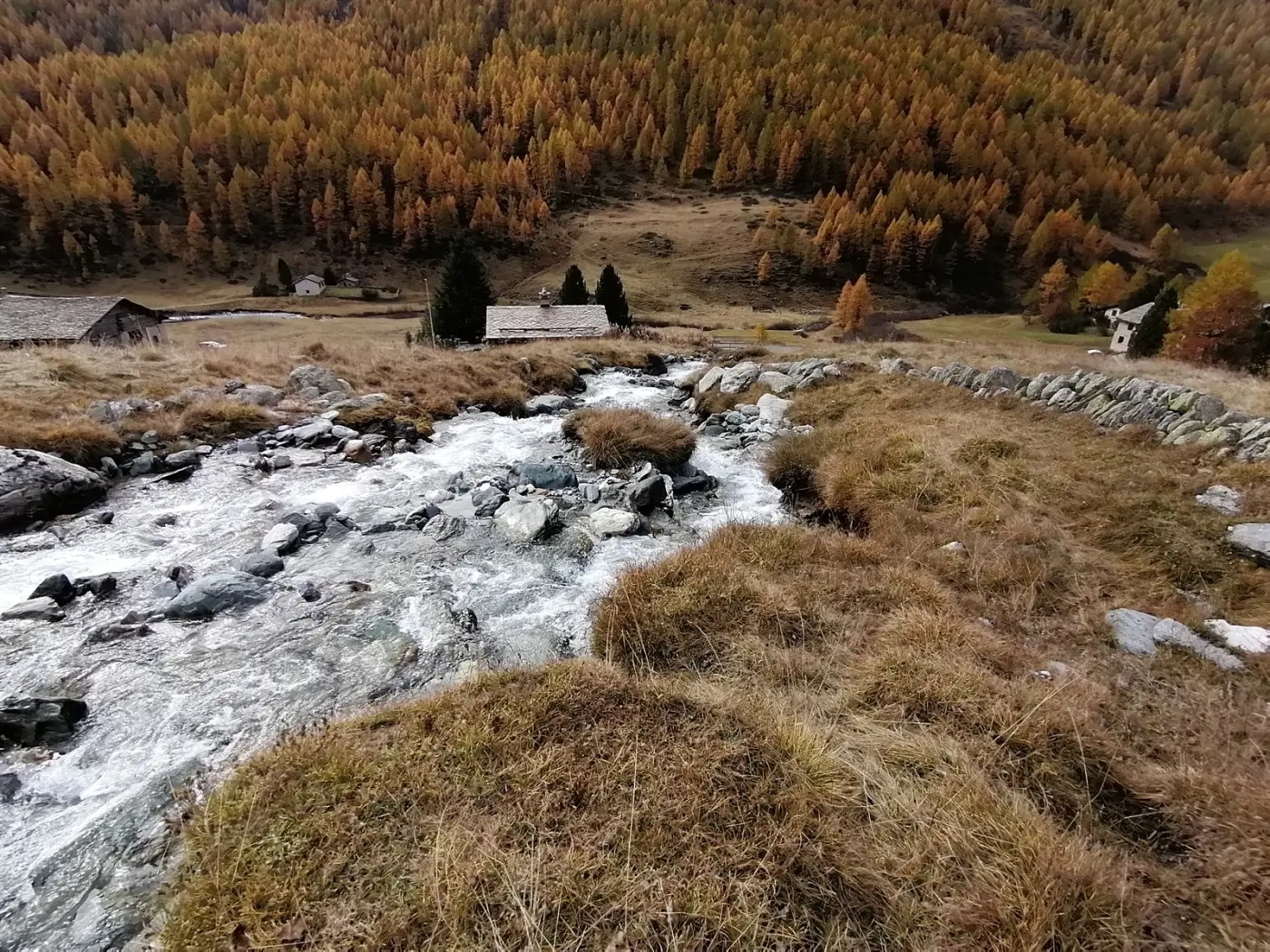 Un ruscello scorre tra le rocce in un paesaggio montano autunnale, con alberi dai colori dorati che circondano un piccolo villaggio. Sullo sfondo si vedono delle baite e un contesto naturale suggestivo.