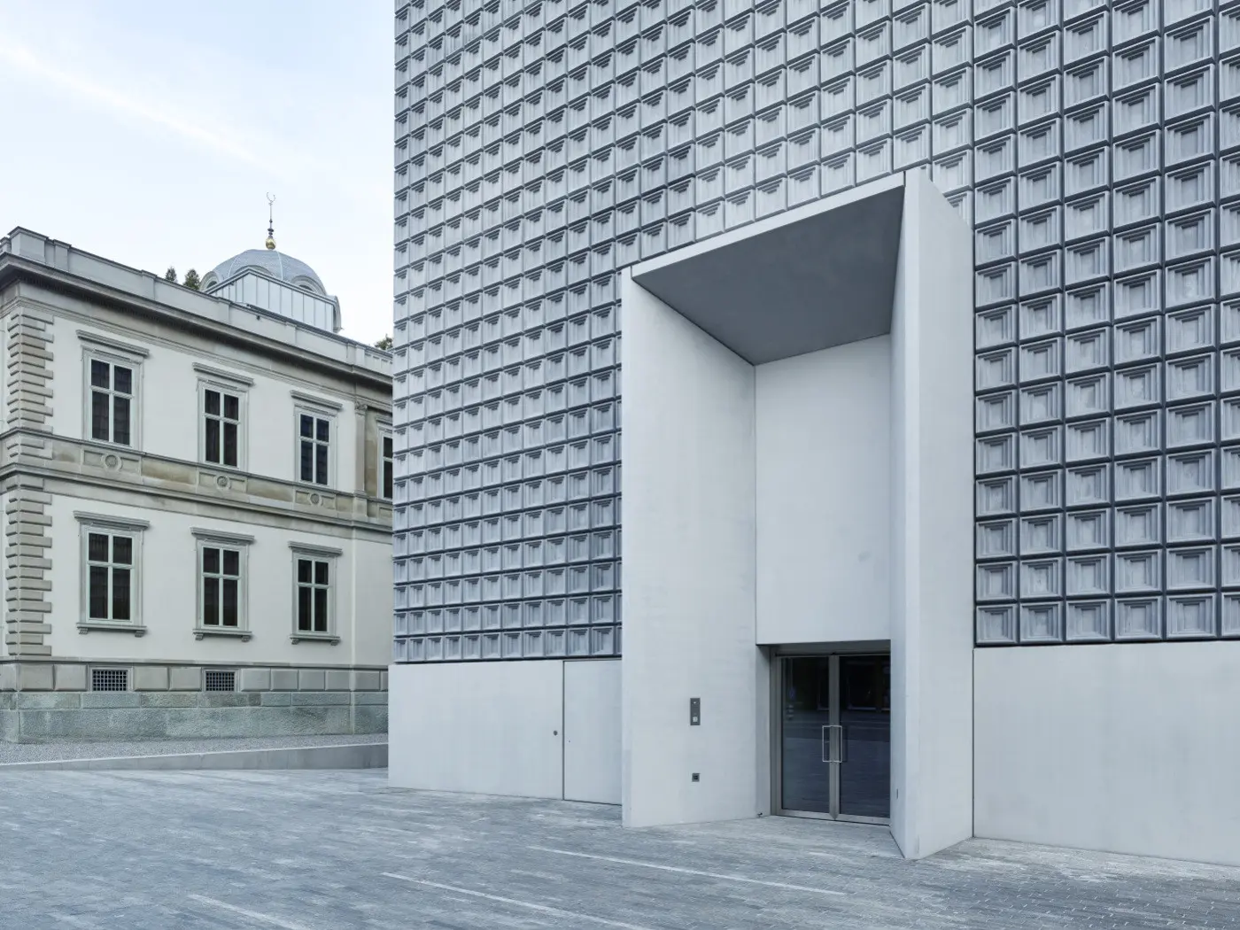Un edificio moderno con una facciata in alluminio e una grande apertura, affiancato da un edificio storico con finestre ornate. Il pavimento è in cemento grigio chiaro.