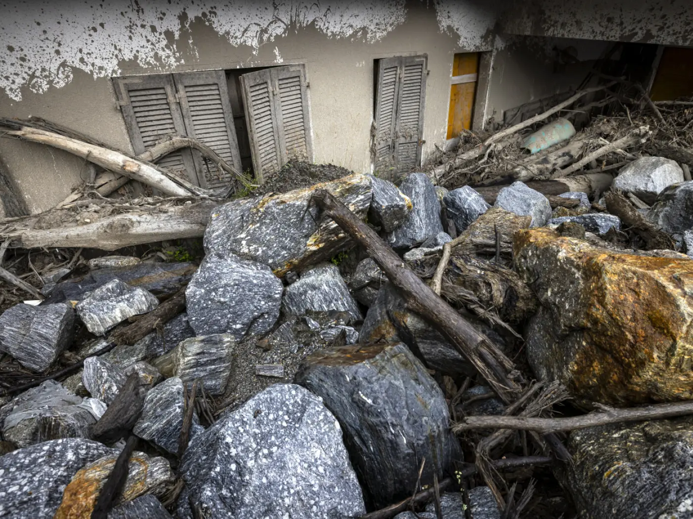 Rocce e detriti di legno accumulati davanti a un edificio gravemente danneggiato, con finestre e persiane visibili sullo sfondo.