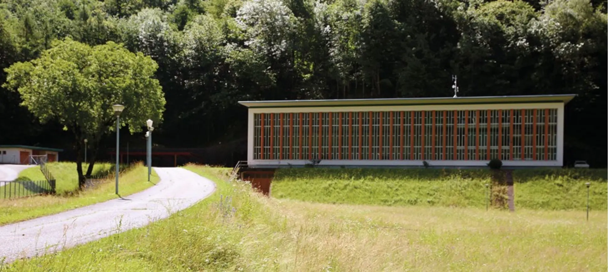 Un edificio moderno con molte finestre colorate si trova su un terreno erboso, circondato da alberi. Una strada sinuosa conduce all'ingresso.