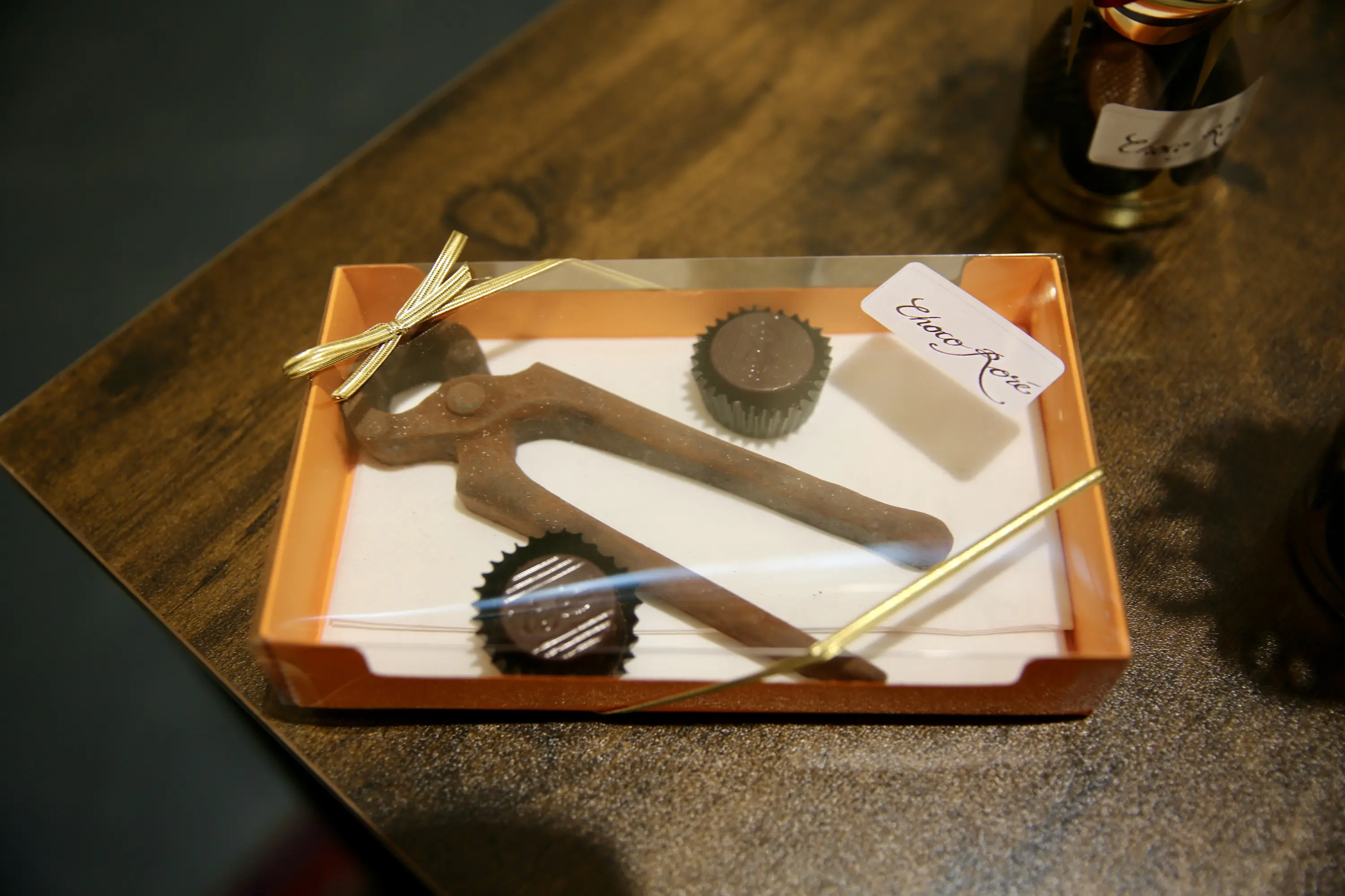 Un regalo in una scatola trasparente con un attrezzo simile a un apribottiglie e due cioccolatini rotondi. La scatola è adornata con un nastro dorato.