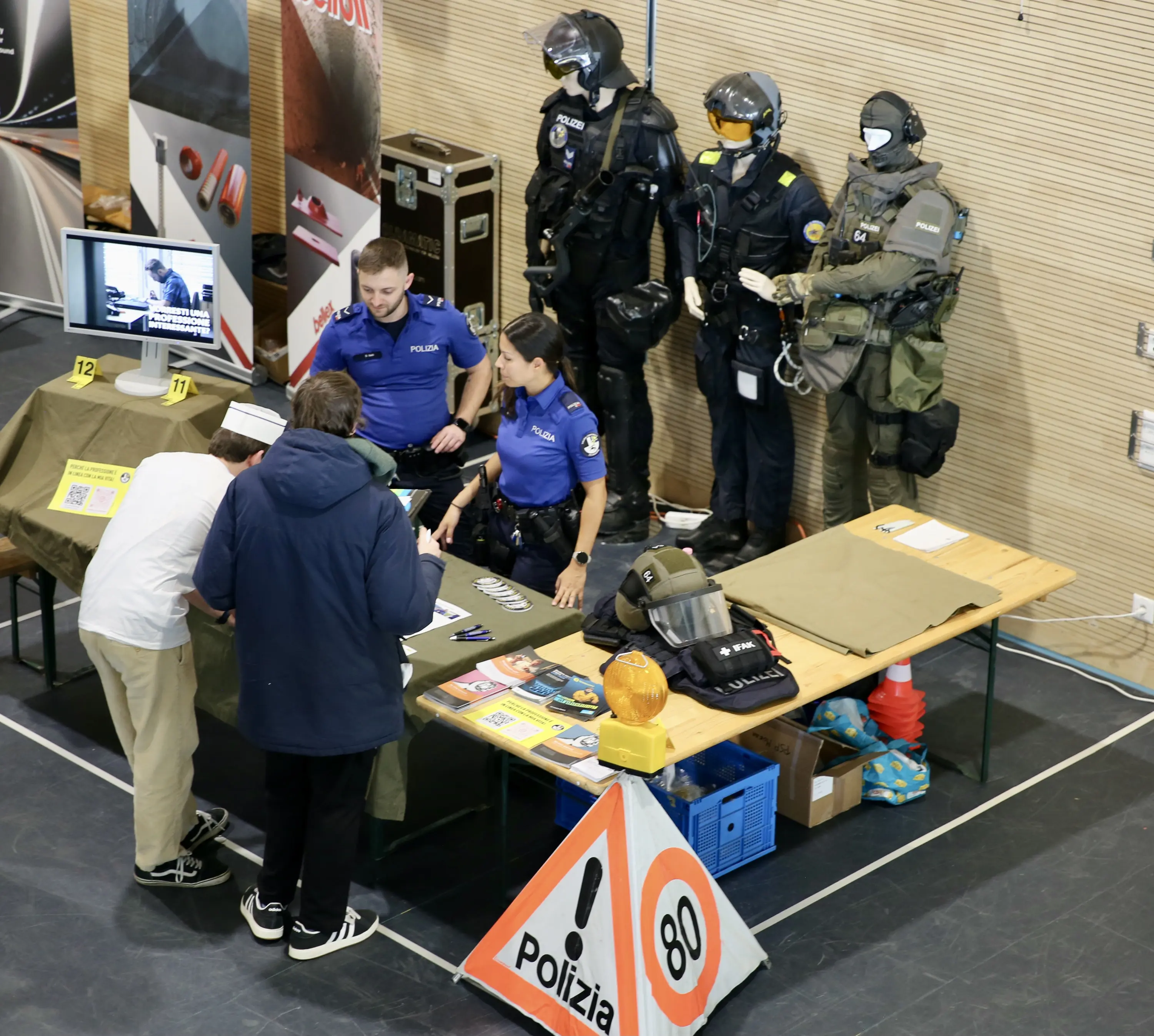 Stand informativo con agenti di polizia in uniforme che interagiscono con i visitatori. Manichini vestiti con equipaggiamento tattico sono esposti. Sul tavolo ci sono materiali informativi e cartelli stradali.