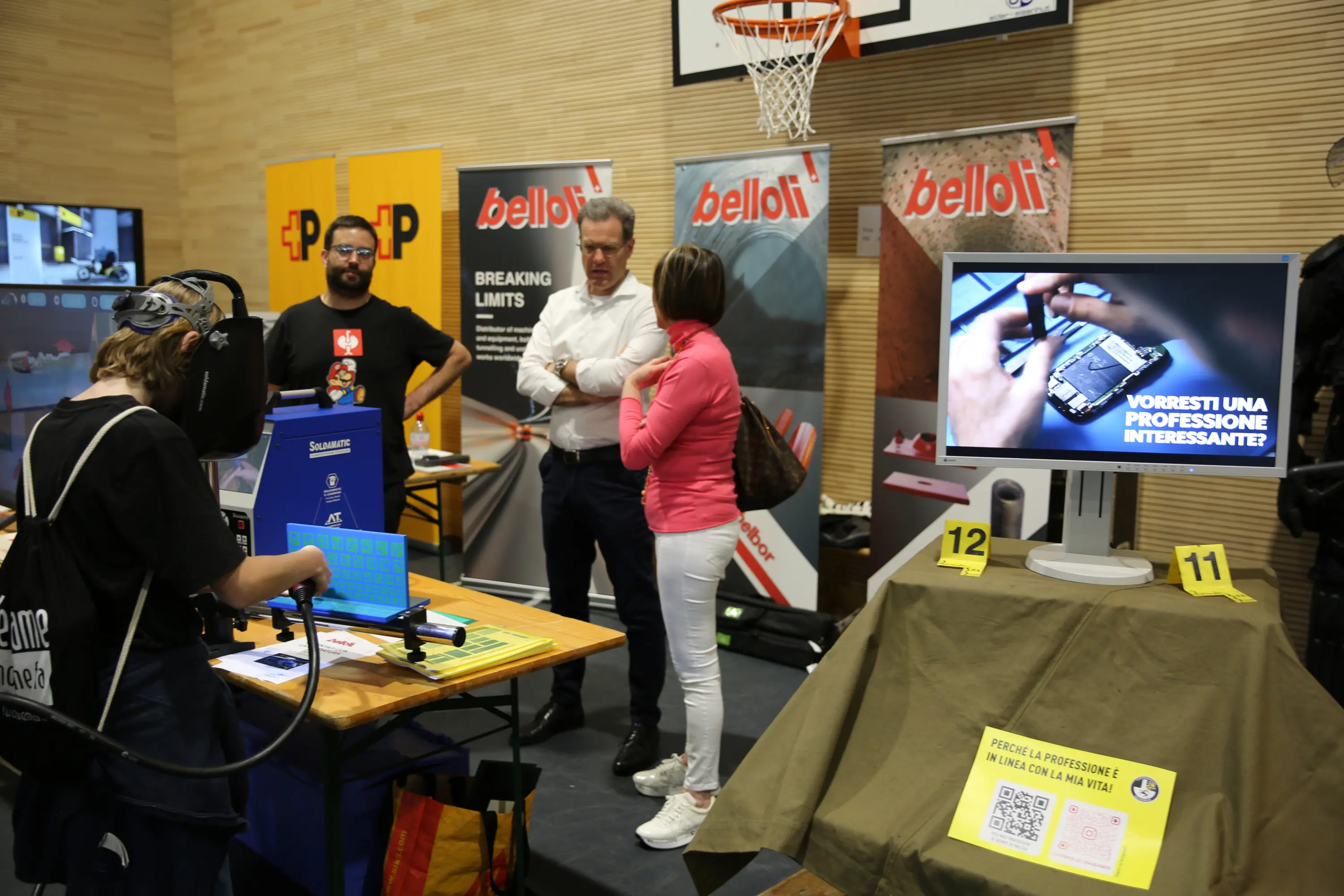 Un evento con diverse stands espositivi. Una persona indossa un visore VR, mentre altre due persone osservano uno schermo che mostra un video. Fondali con scritte pubblicitarie e un canestro da basket visibile sullo sfondo.