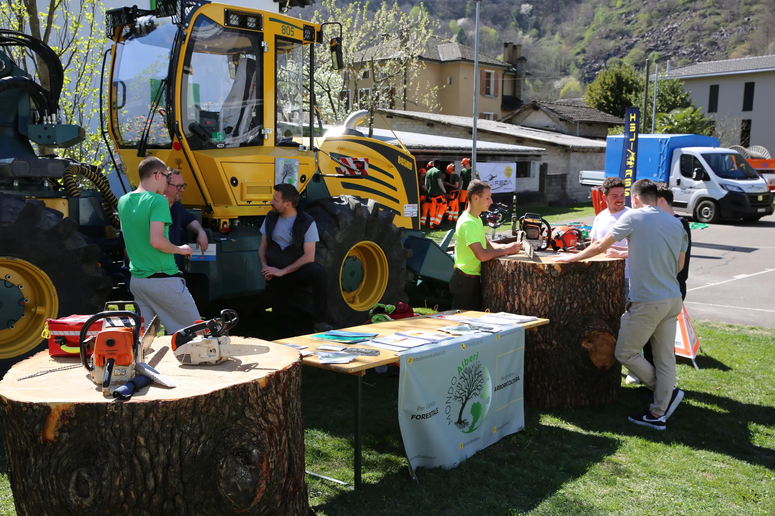 Un gruppo di persone interagisce vicino a un grande veicolo agricolo, con tronchi d'albero e attrezzature da lavoro, come seghe, disposti su un tavolo. In lontananza si vedono altri veicoli e edifici.