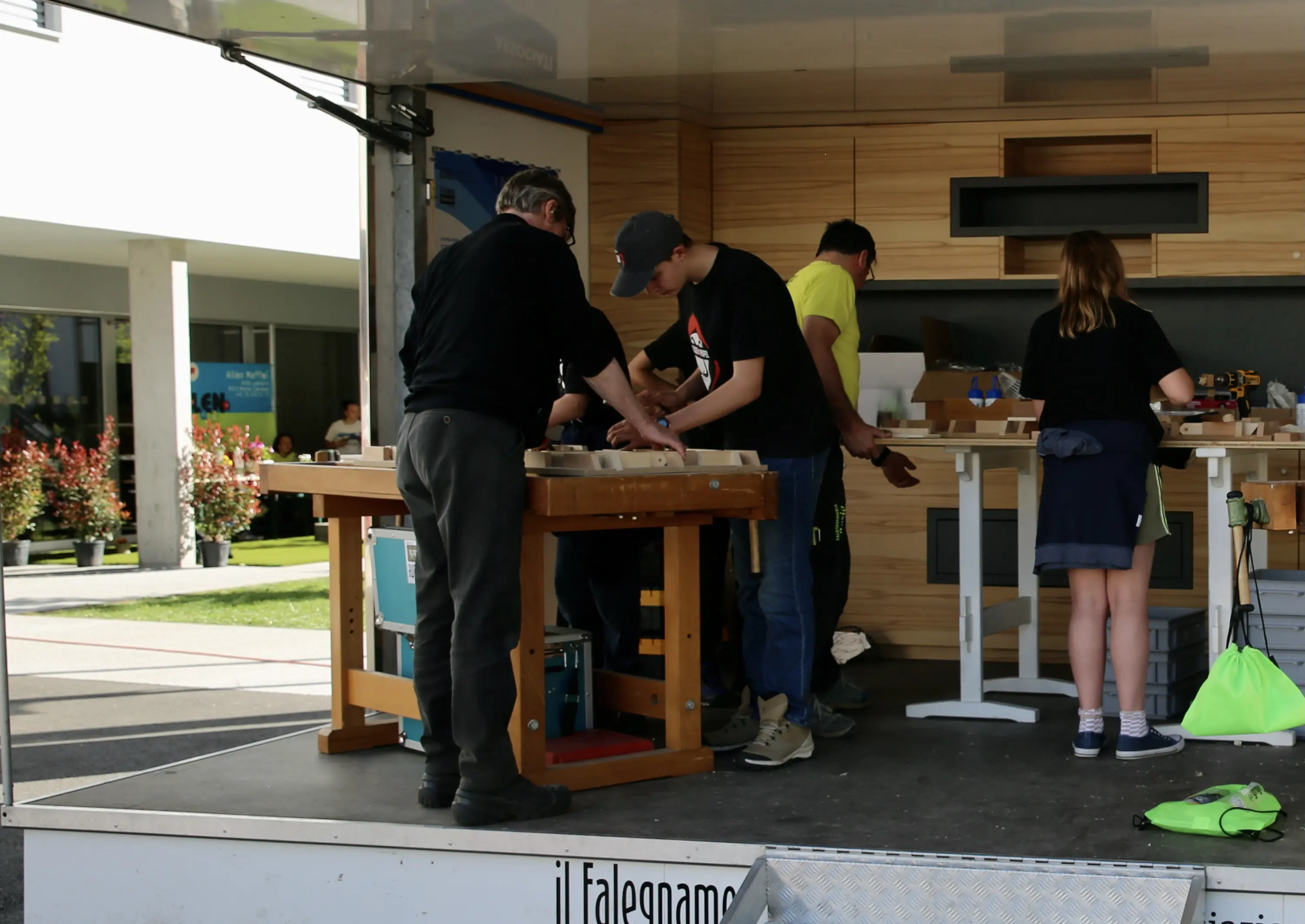 Un gruppo di persone sta lavorando a un banco da lavoro all'aperto, probabilmente durante un laboratorio di falegnameria. Alcuni partecipanti stanno tagliando o assemblando materiali, mentre altri osservano e assistono. Si nota un ambiente di collaborazione e apprendimento.