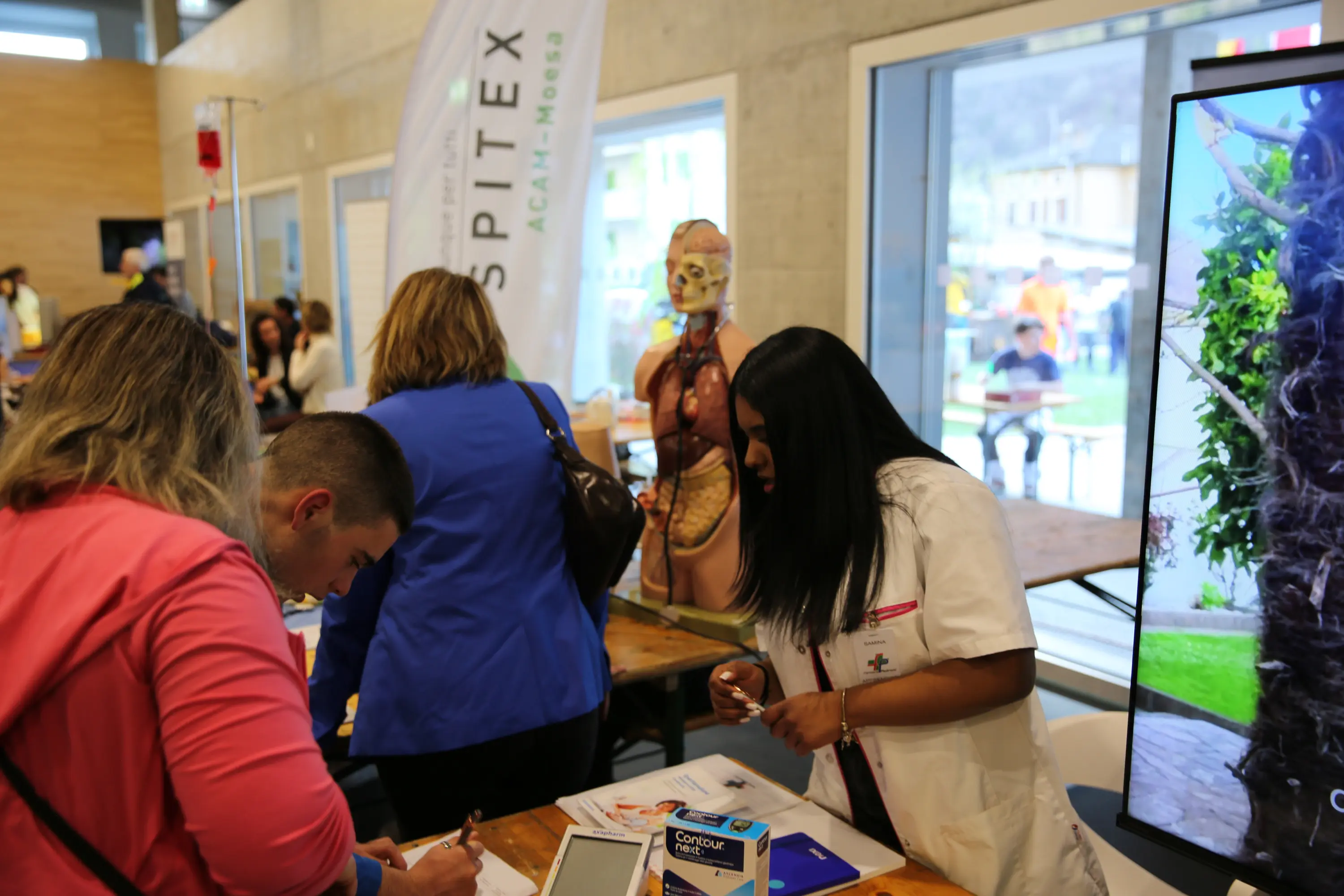 Una fiera con persone che interagiscono a uno stand. Una donna in camice bianco sta parlando con un visitatore, mentre un manichino anatomico è visibile sullo sfondo. Un grande schermo mostra immagini relative all'evento.