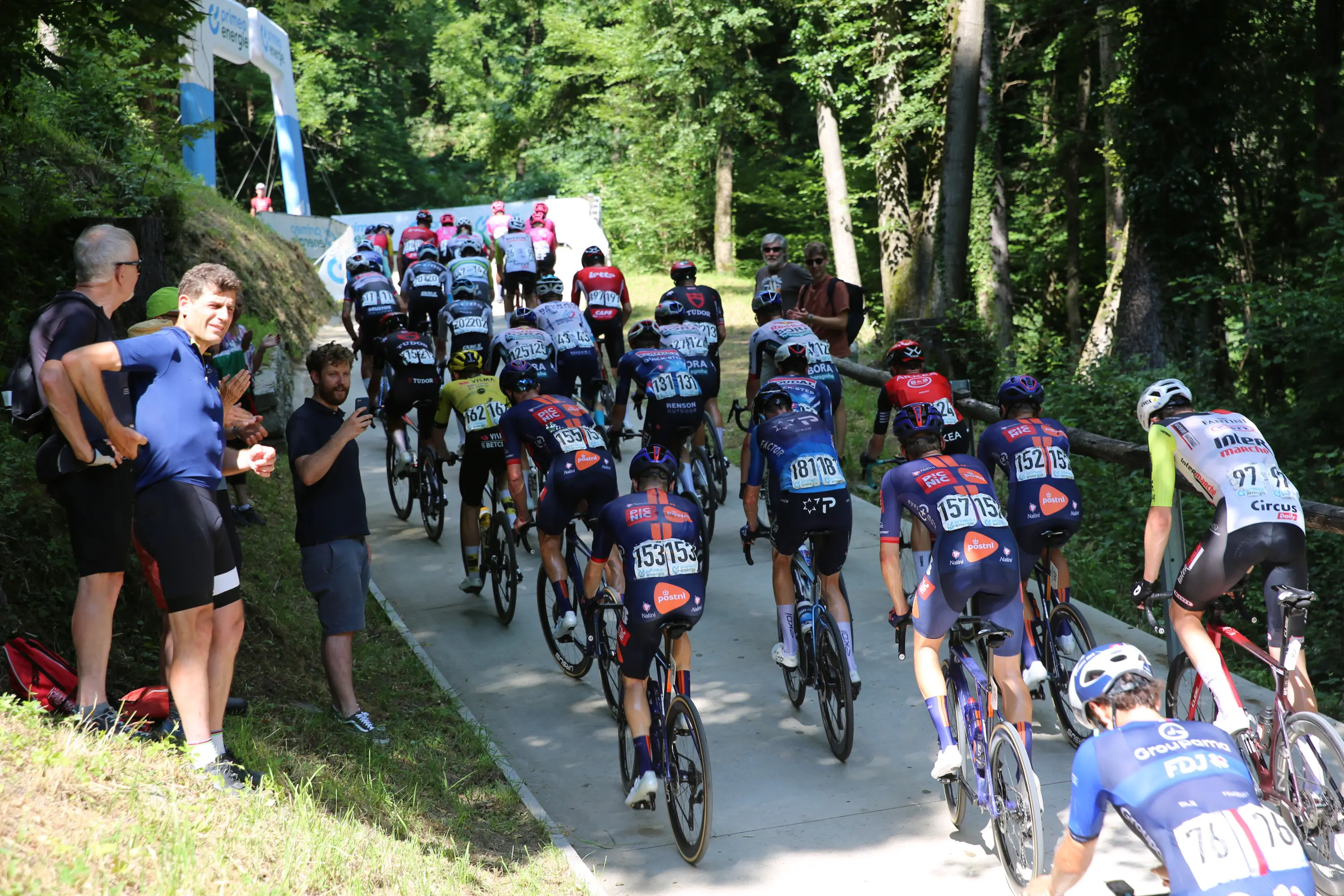 Gruppo di ciclisti in gara su una strada sterrata circondata da alberi. Alcuni spettatori osservano dalla sinistra. I ciclisti indossano varie maglie da competizione.