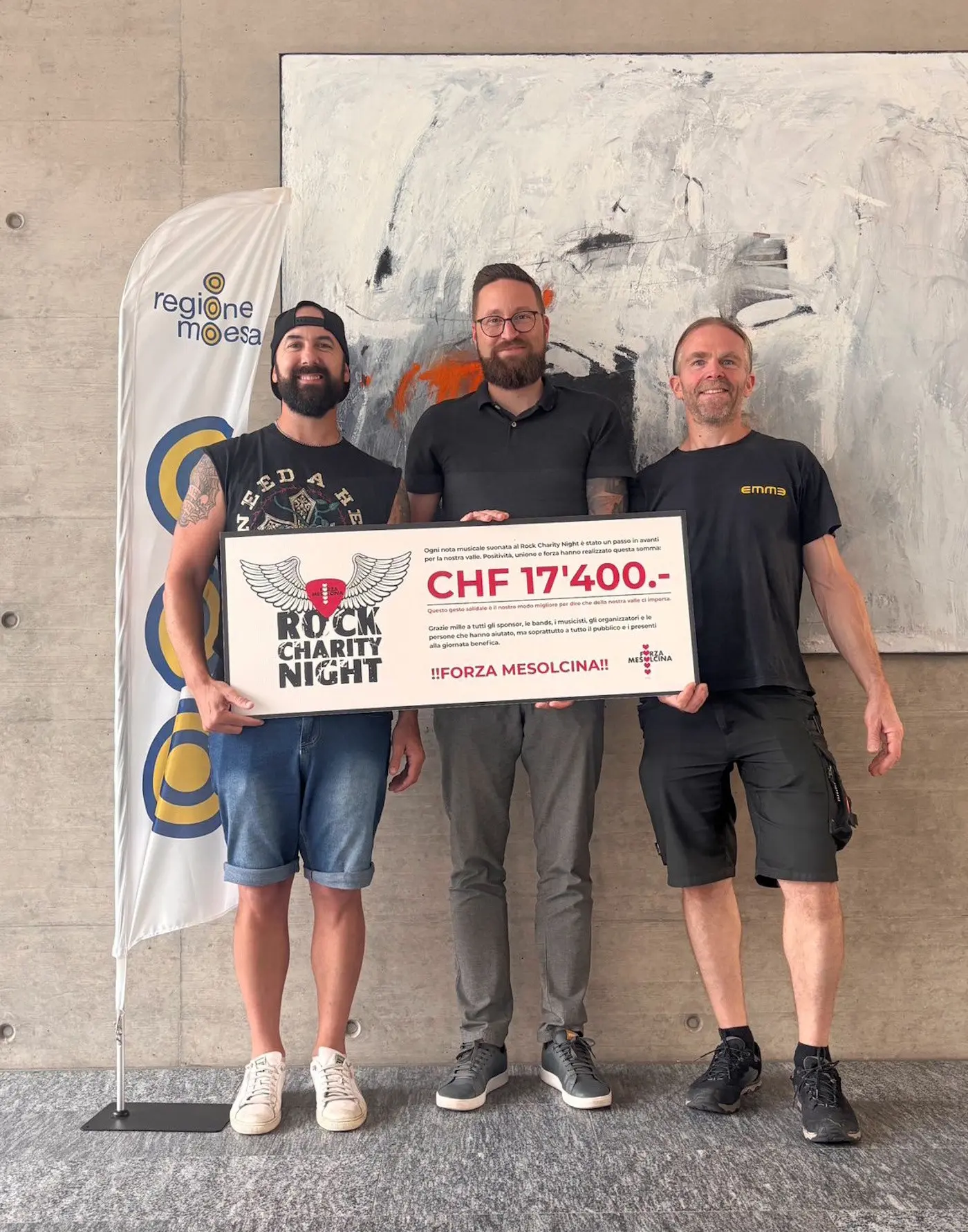 Tre uomini sorridenti posano insieme, tenendo un grande assegno con l'importo di CHF 17'400. Sullo sfondo, una parete grigia e una bandiera con il logo "Regione Moesa".