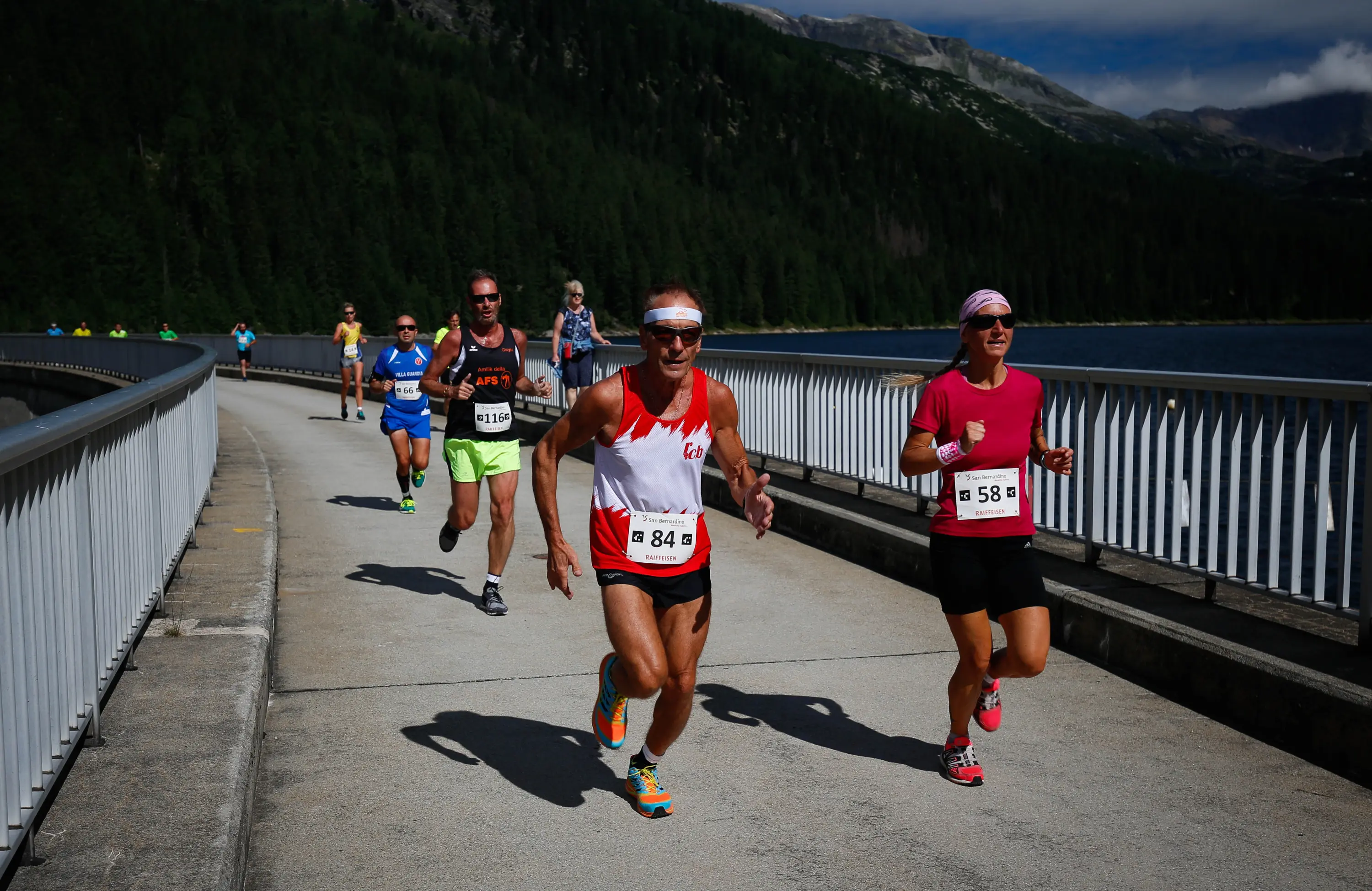 Gruppo di corridori in competizione su una pista lungo un lago, circondati da montagne verdi. Un corridore indossa una maglietta rossa e bianca con il numero 84, mentre un'altra corridore ha una maglietta fucsia con il numero 58. Sole splendente e cielo blu.