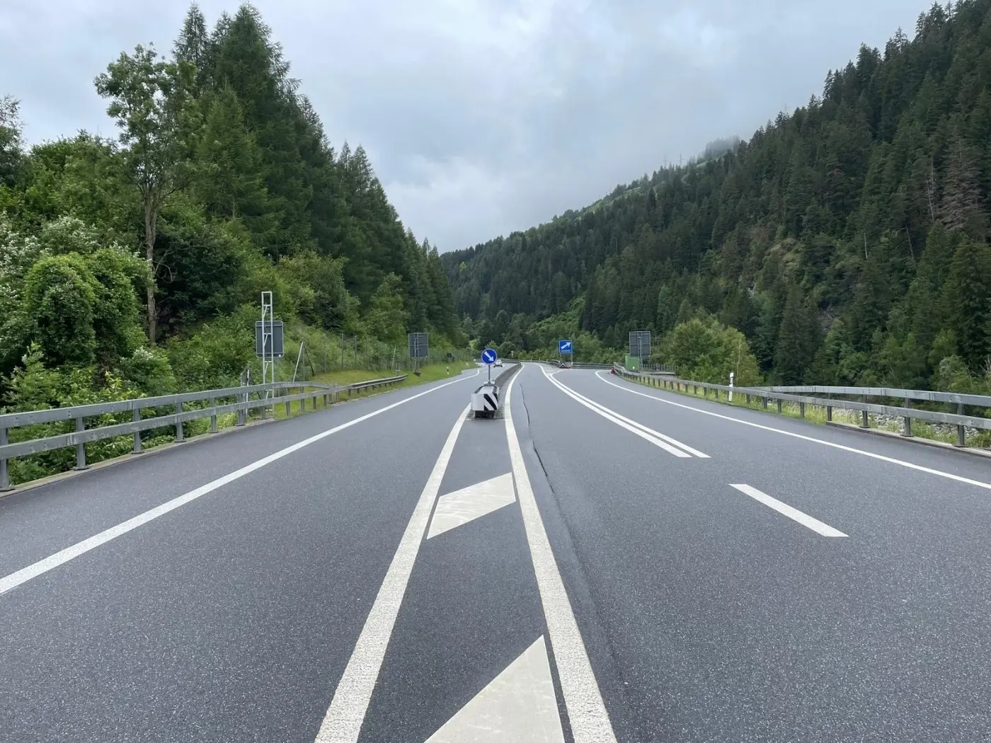 Una strada a doppia corsia che si snoda tra alberi verdi, con una carreggiata bagnata e segnaletica stradale visibile. Il cielo è nuvoloso e la scena appare tranquilla.