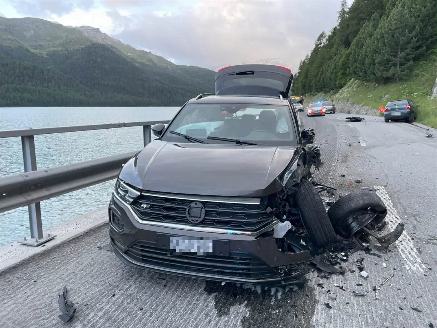 Un'auto nera danneggiata sul ciglio di una strada, con il lato anteriore gravemente incidentato e una gomma mancante. Sullo sfondo, un lago e montagne.