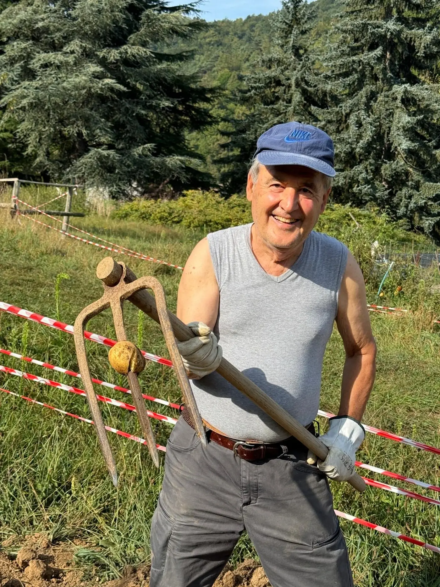 Un uomo sorridente indossa una maglietta senza maniche e un cappellino mentre tiene un forcone da giardinaggio e una palla di terra in un campo. Sullo sfondo si vedono alberi e recinzioni.