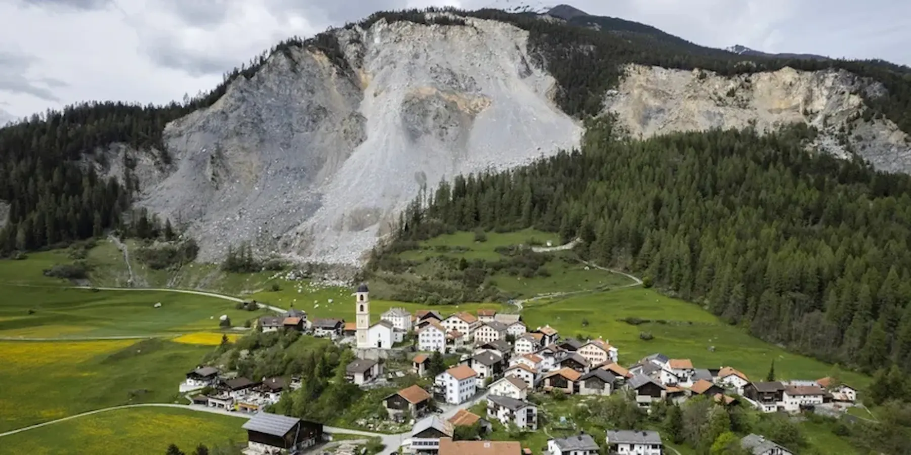 Un villaggio pittoresco immerso nei boschi, con case tradizionali e una chiesa al centro. Sullo sfondo, una grande montagna rocciosa con segni di frane e vegetazione. Aggiungi campi verdi e gialli in primo piano.