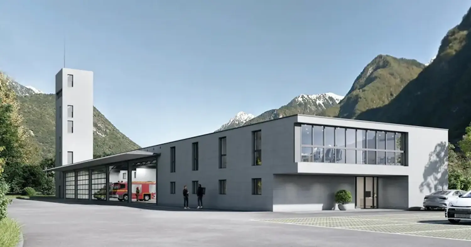 Edificio moderno e minimalista, con una torre slanciata, situato in un paesaggio montano. La struttura ha ampie finestre e un'area garage per veicoli, con un camion dei pompieri visibile all'interno. Due persone sono per strada davanti all'edificio.