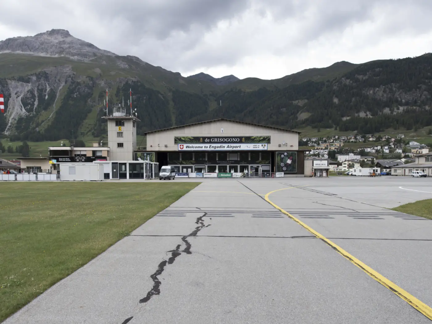L'aeroporto di Samedan è visibile con la sua facciata principale e il logo "Welcome to Engadin Airport". Il panorama montano circostante è verde con nuvole nel cielo. La pista è in primo piano.