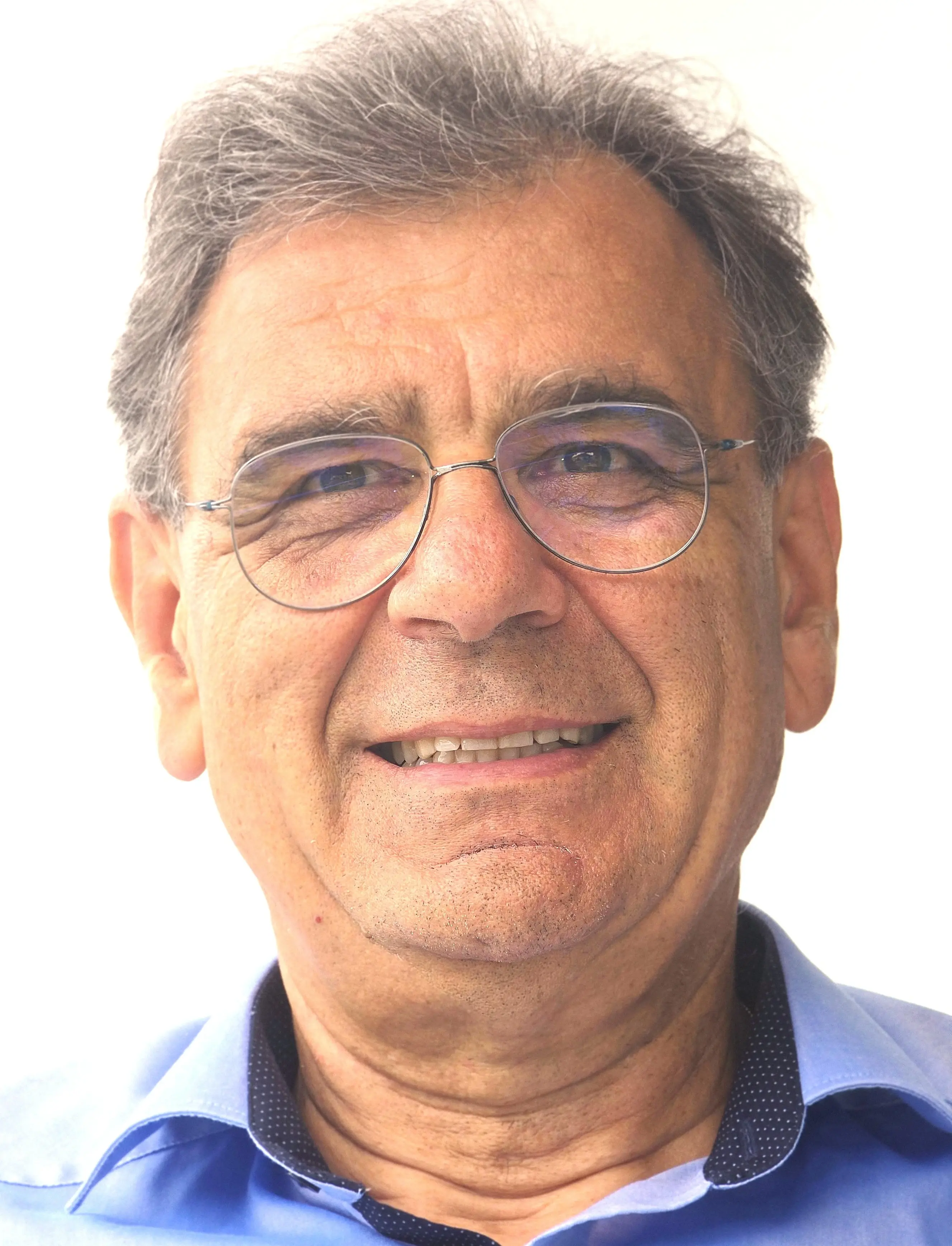 Uomo sorridente con capelli grigi e occhiali, indossa una camicia blu e ha uno sfondo chiaro.