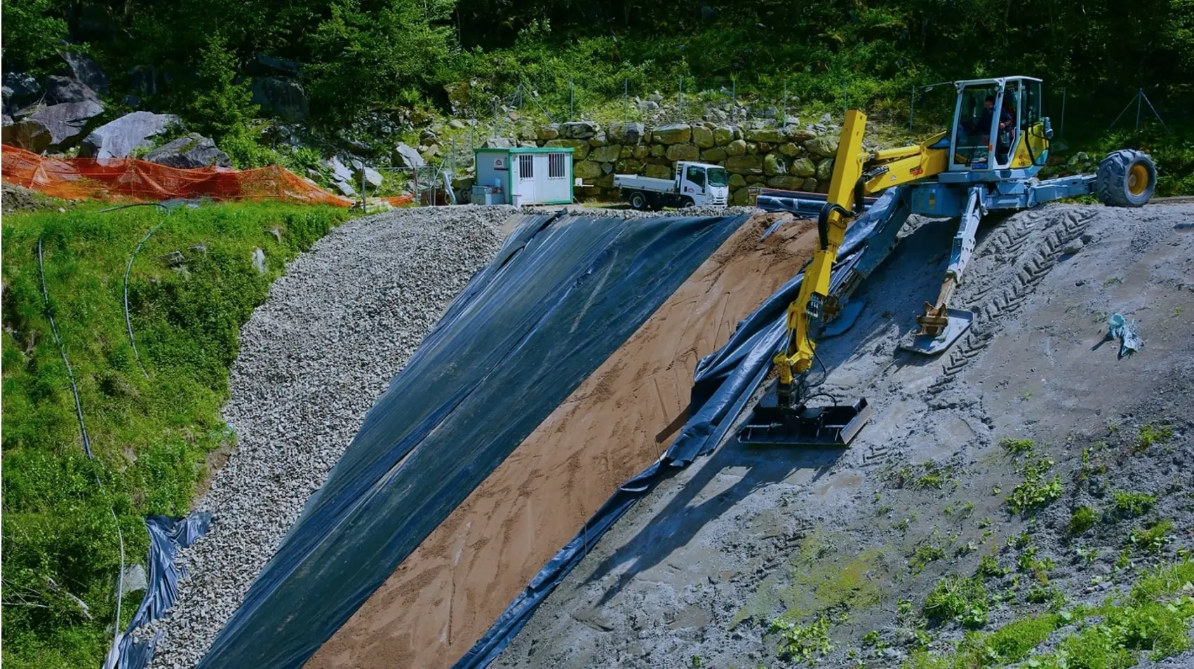 Macchinari per la costruzione e il rinforzo del terreno, con una pala meccanica impegnata nel posizionamento di materiali su una scarpata. Sullo sfondo si vedono un camion e una cabina di cantiere.