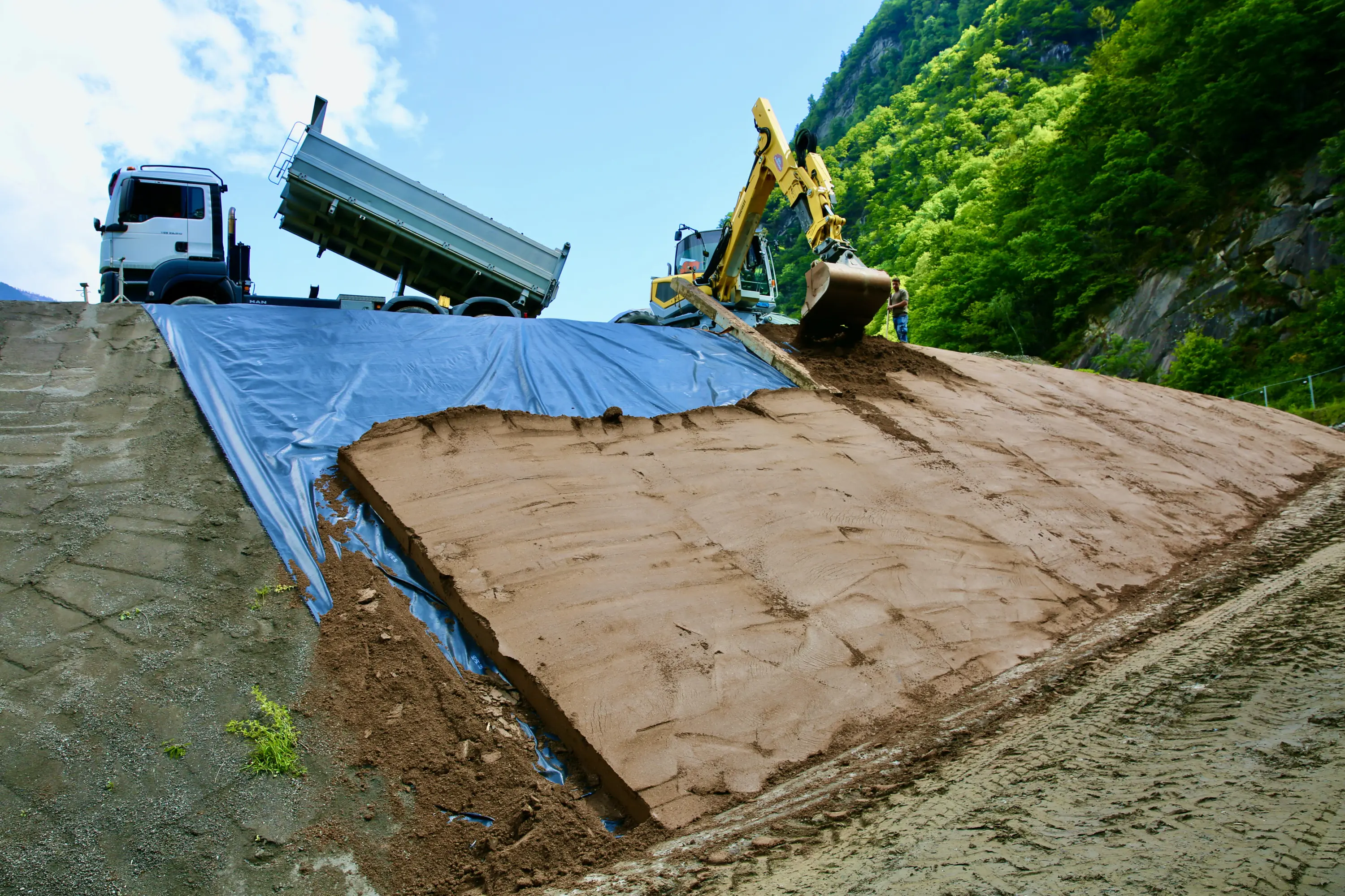 Un escavatore sta spostando terreno su una pendenza, mentre un camion è parcheggiato in alto. Una tela blu copre la parte superiore della pendenza. L'ambiente circostante è collinare e verdeggiante.