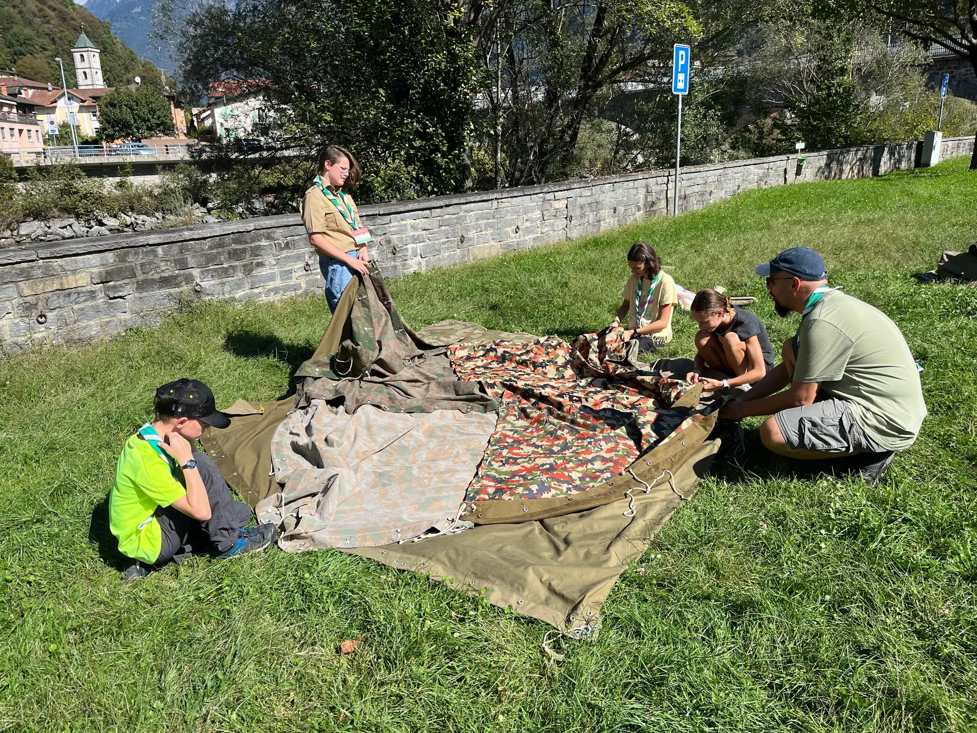 Un gruppo di quattro giovani all'aperto sta montando una tenda su un prato. Due ragazzi siedono sul terreno mentre due ragazze stanno in piedi, tutte sono concentrate sull'operazione. Sullo sfondo si vede una strada e delle montagne.