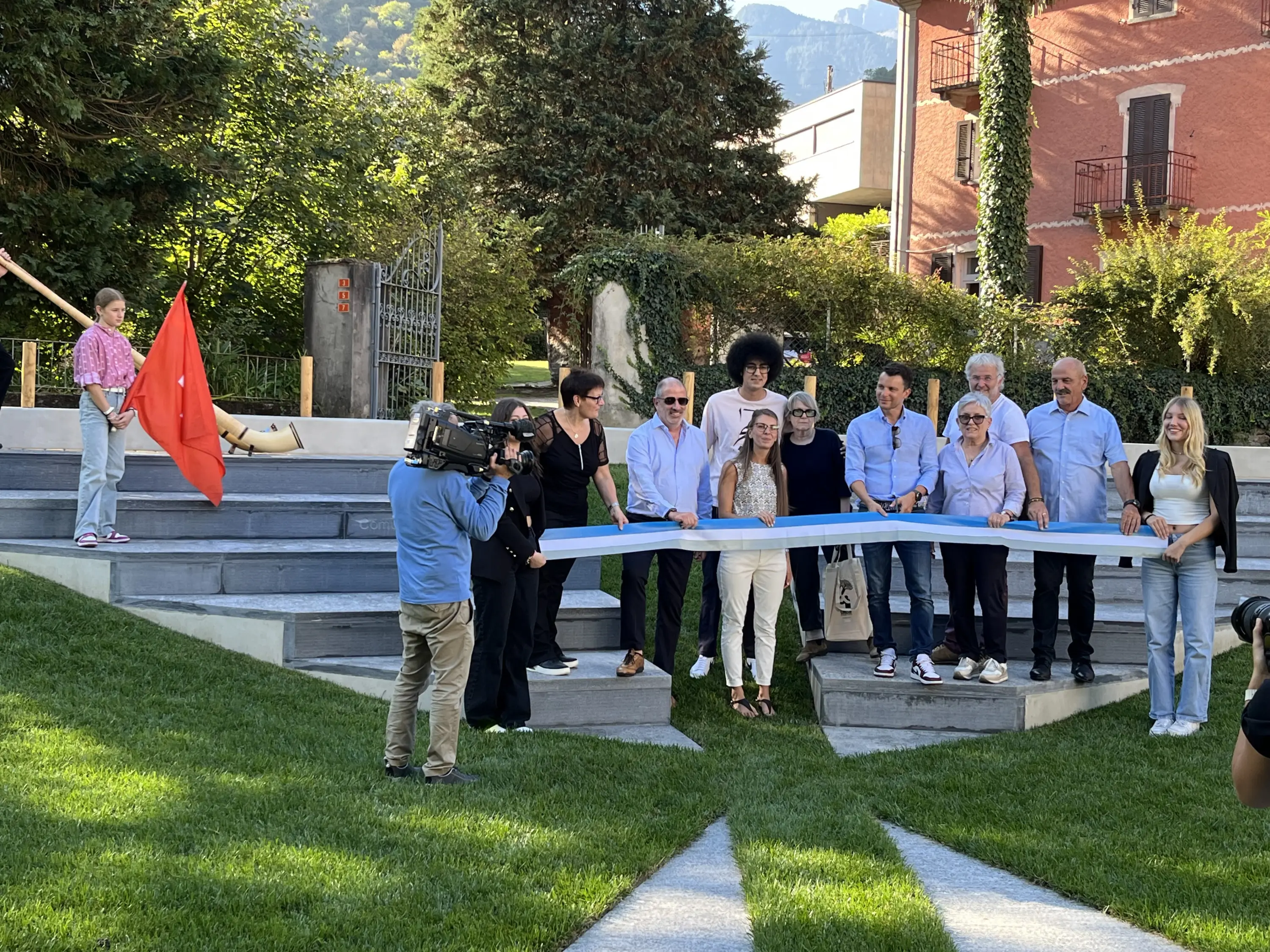 Cerimonia di inaugurazione con un gruppo di persone che posano davanti a un nastro da tagliare. Alcuni partecipanti tengono una bandiera rossa. Una cameraman riprende l'evento in un ambiente all'aperto con edifici e vegetazione sullo sfondo.