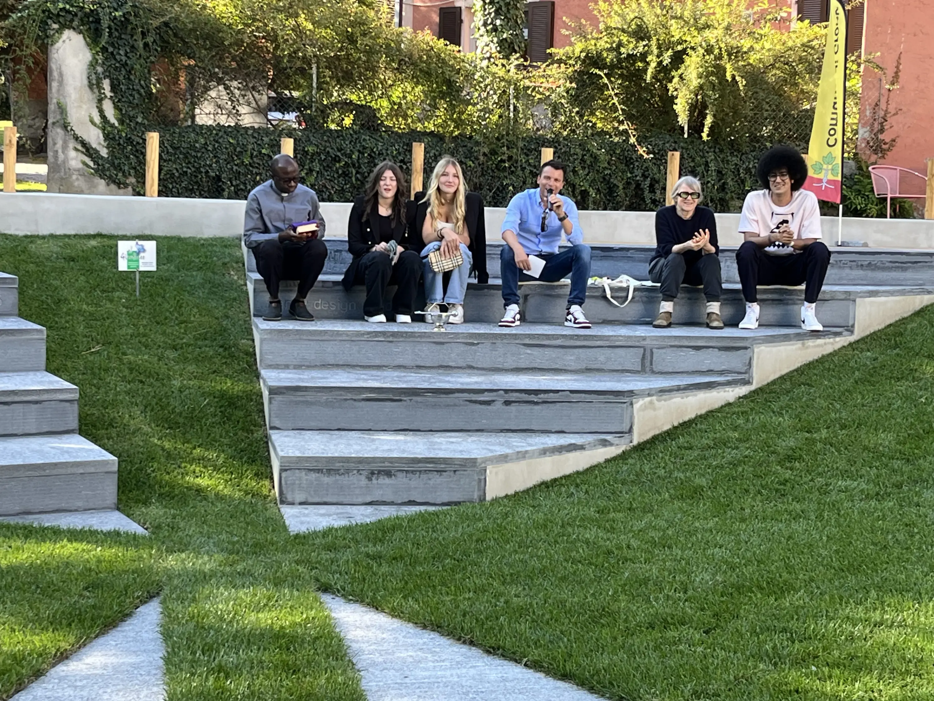 Un gruppo di sei persone sedute su gradini di un'area verde, immerse nella conversazione e gustando cibo. Sullo sfondo, piante e architettura moderna.