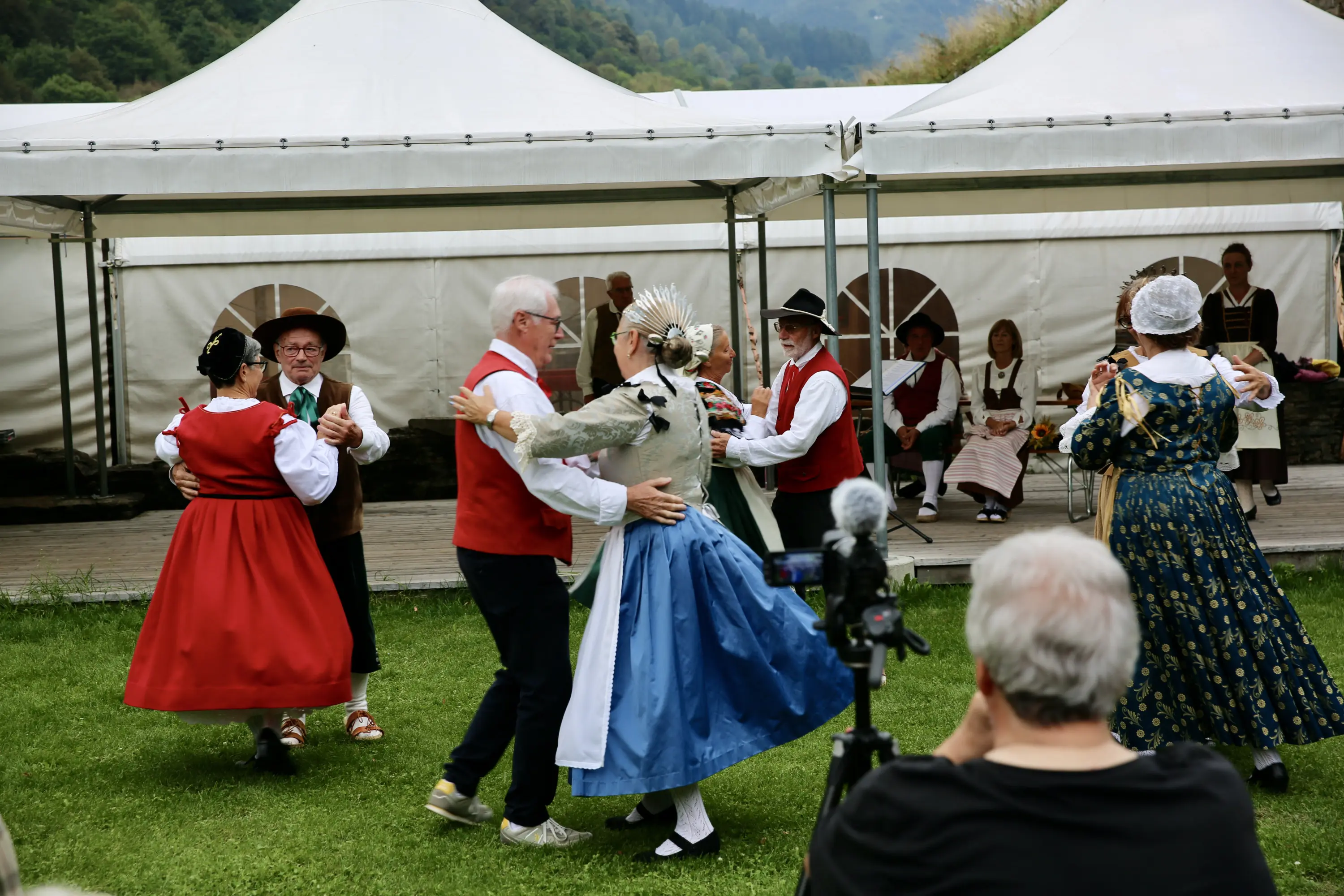 Persone in abiti tradizionali danzano insieme in un festival folkloristico, con una selezione di tende bianche sullo sfondo e spettatori che osservano.