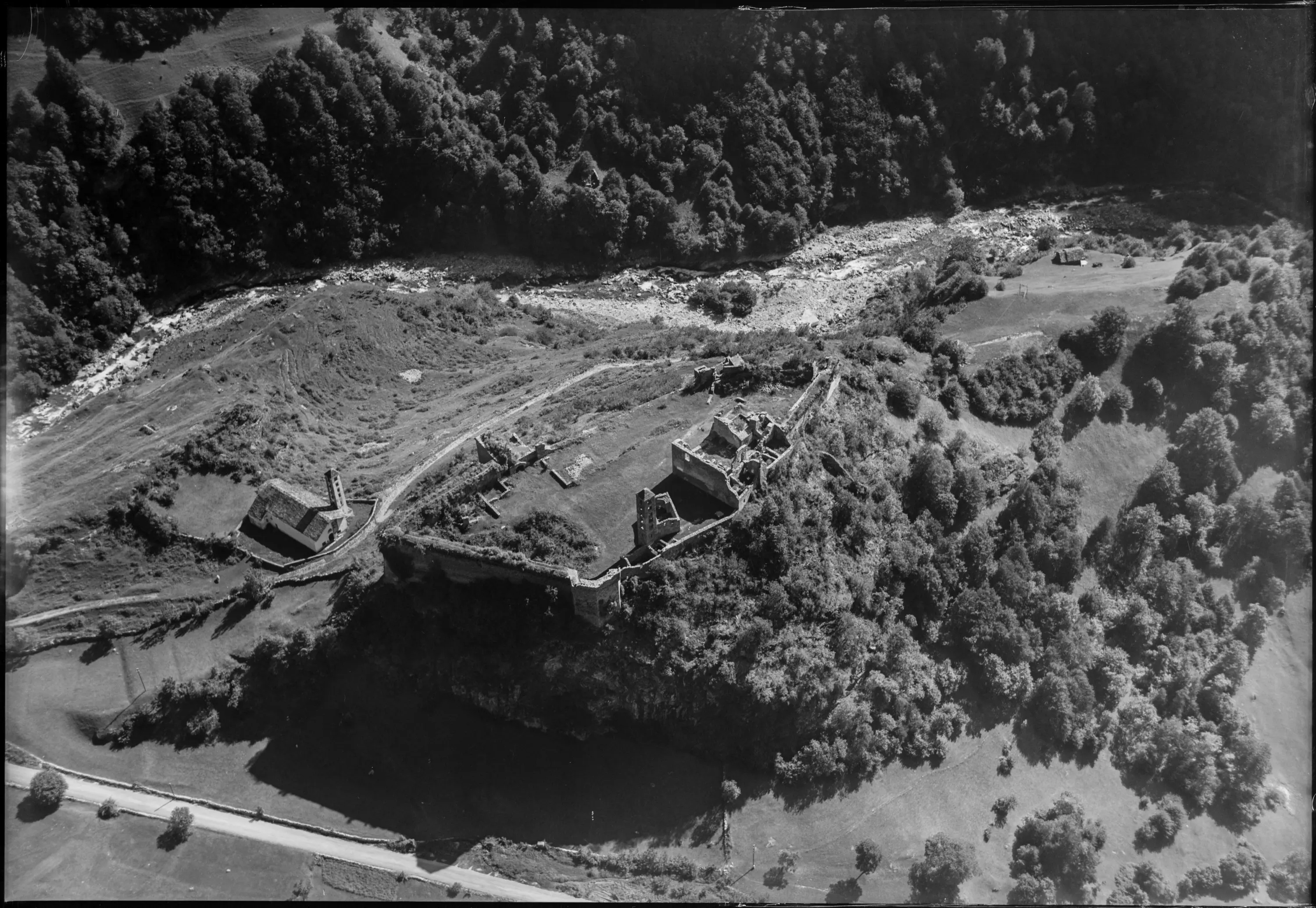 Vista aerea di una struttura fortificata situata su una collina, circondata da vegetazione e un fiume che scorre nelle vicinanze. Il paesaggio circostante è verde e collinare.