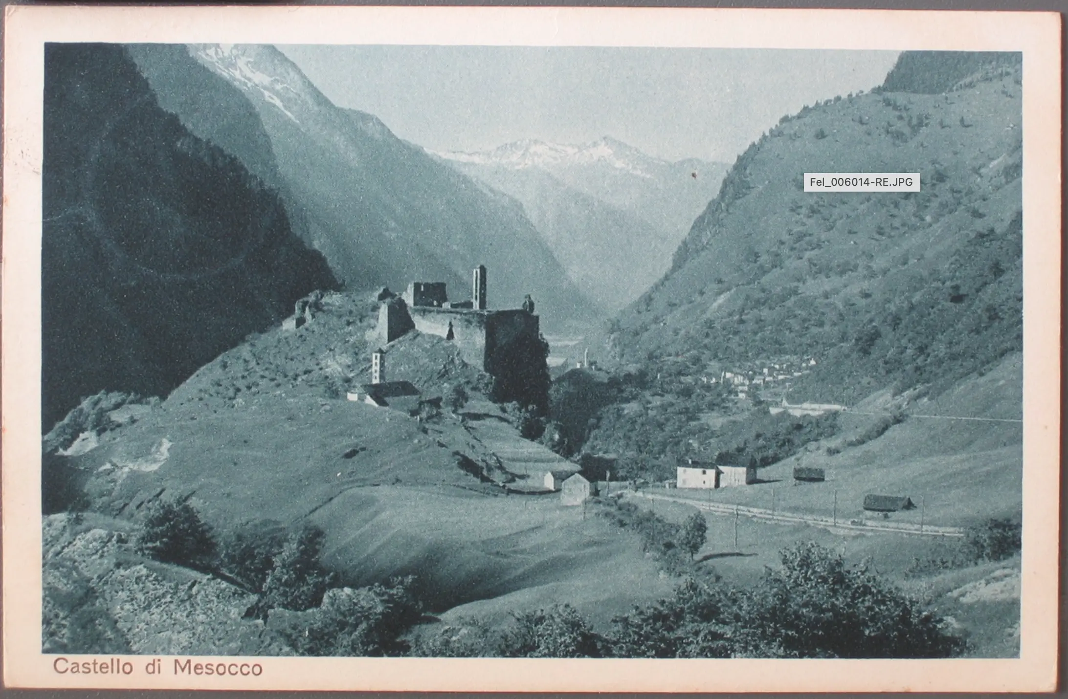 Veduta del Castello di Mesocco, situato in una valle circondata da montagne. Il castello è parzialmente rovinato e circondato da una vegetazione verde, con alcune costruzioni nel fondovalle.