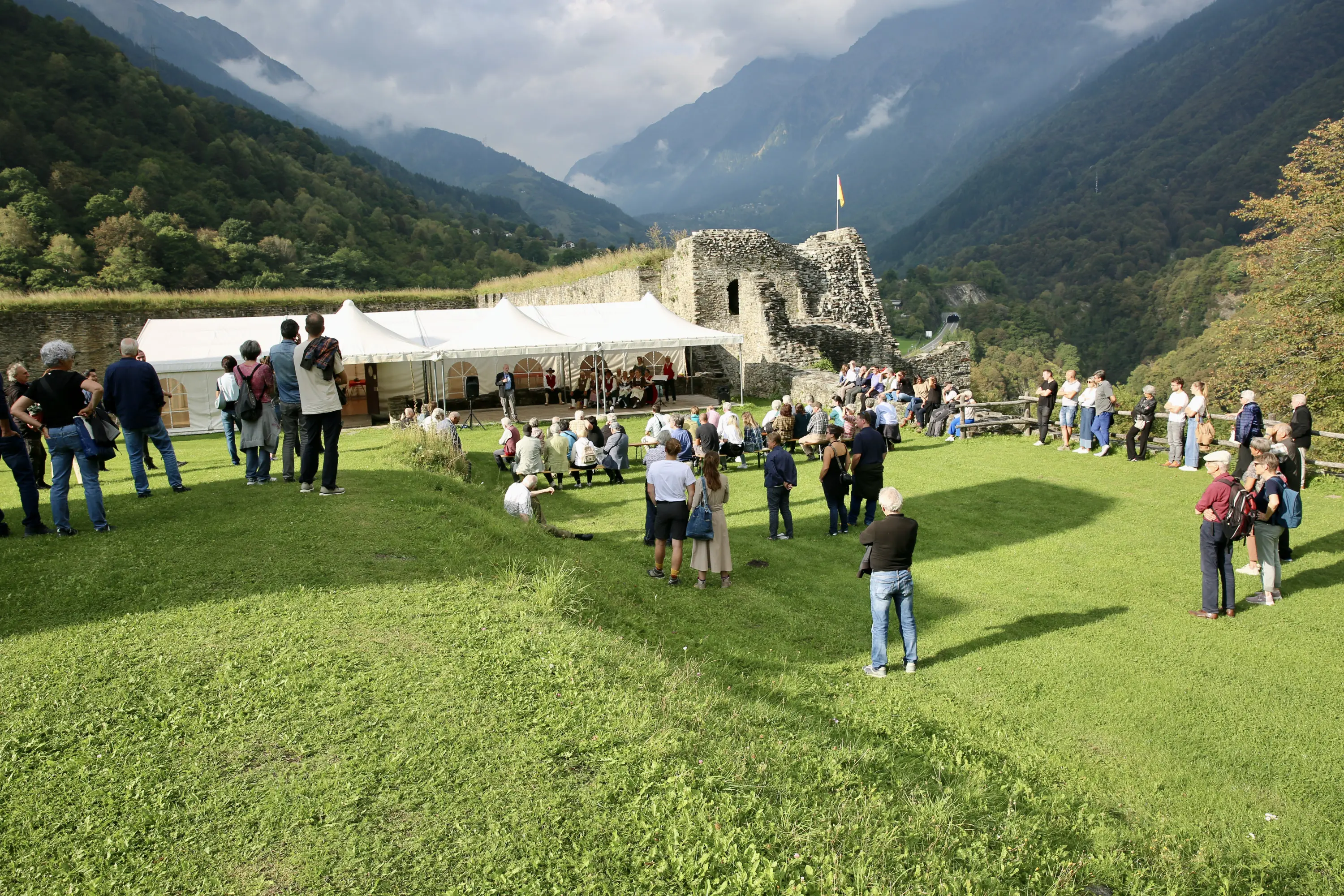 Un evento all'aperto si svolge in un paesaggio montano. Persone si radunano intorno a un palco allestito sotto un tendone. Sullo sfondo, resti di un'antica costruzione e alberi verdi. Le montagne si ergono maestose sotto un cielo nuvoloso.