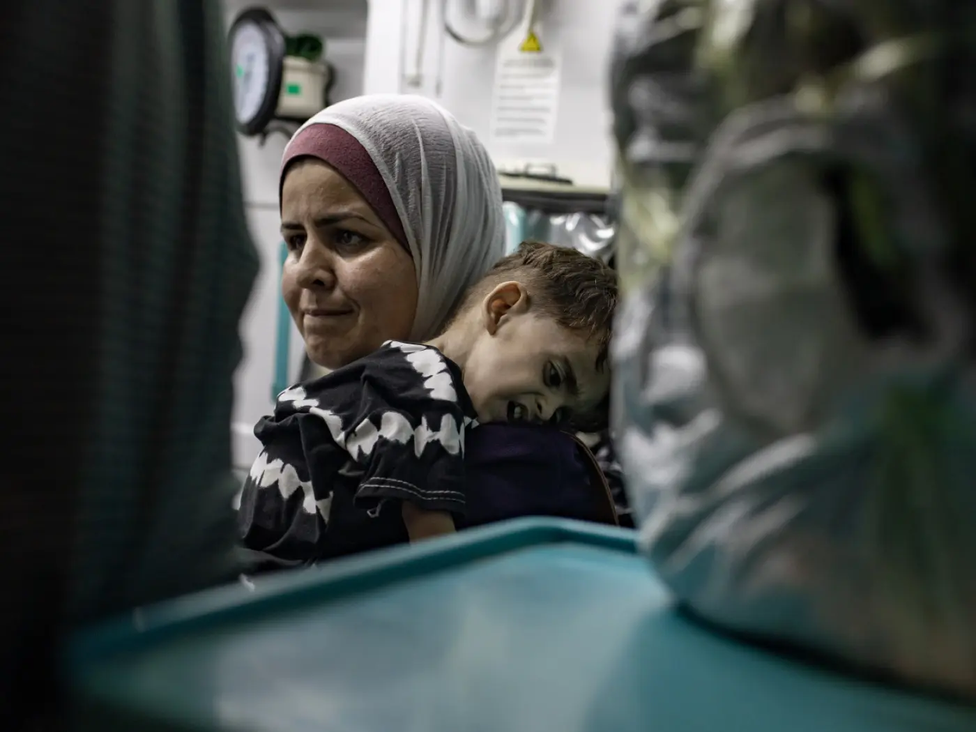 Una donna con un velo guarda in lontananza mentre tiene in braccio un bambino. Il bambino sembra stanco e appoggiato contro di lei. Sullo sfondo ci sono oggetti e attrezzature mediche.