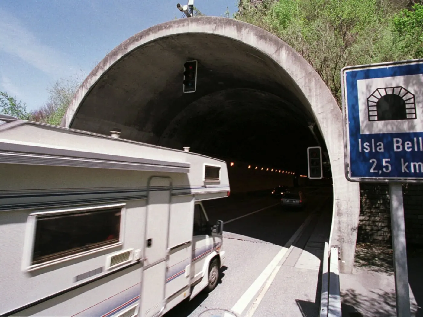 Un camper bianco entra in un tunnel, con semafori rossi e blu visibili. Un cartello indica la direzione verso Isla Bella a 2,5 km.