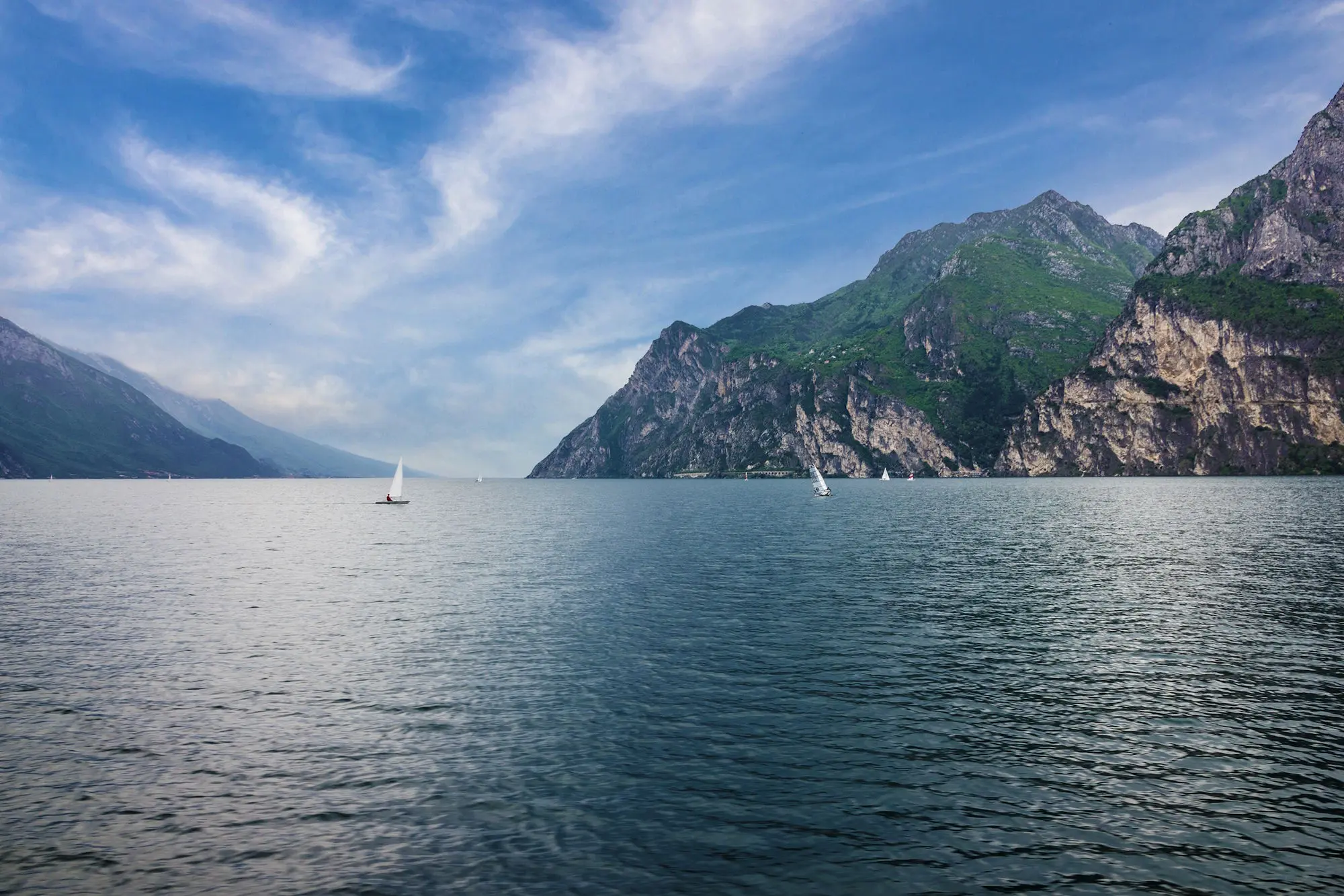 Lago circondato da imponenti montagne verdi, con piccole barche a vela che navigano sull'acqua calma sotto un cielo blu con nuvole leggere.