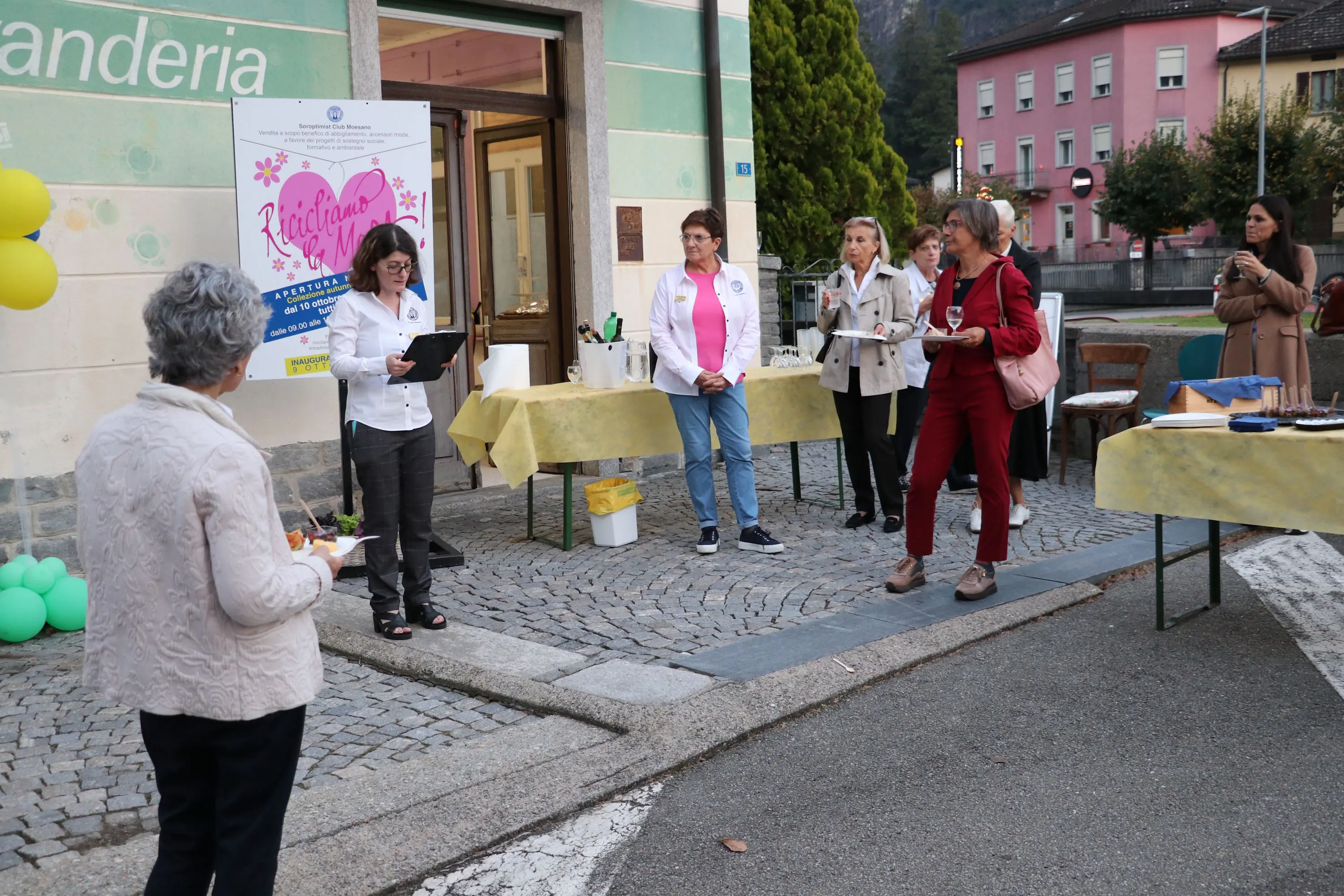 Un gruppo di donne si riunisce all'esterno di un edificio per un evento. Una donna con una lavagna parla al pubblico, mentre altre ascoltano e registrano. Sono presenti decorazioni e alcuni tavoli imbanditi.