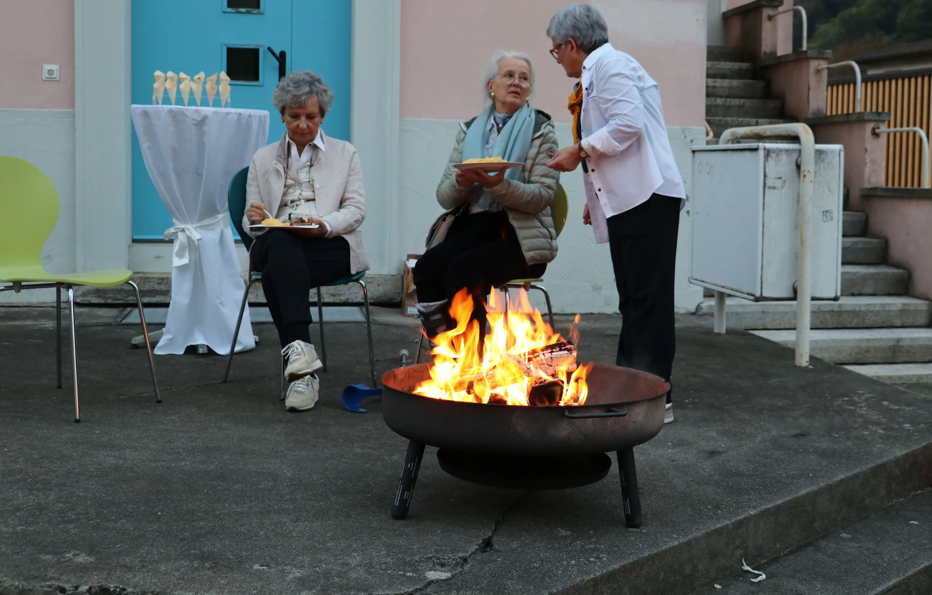 Un falò acceso su un patio, con tre donne sedute nei dintorni. Due donne chiacchierano mentre una terza tiene un piatto. Sullo sfondo, una porta blu e una tavola imbandita con decorazioni.