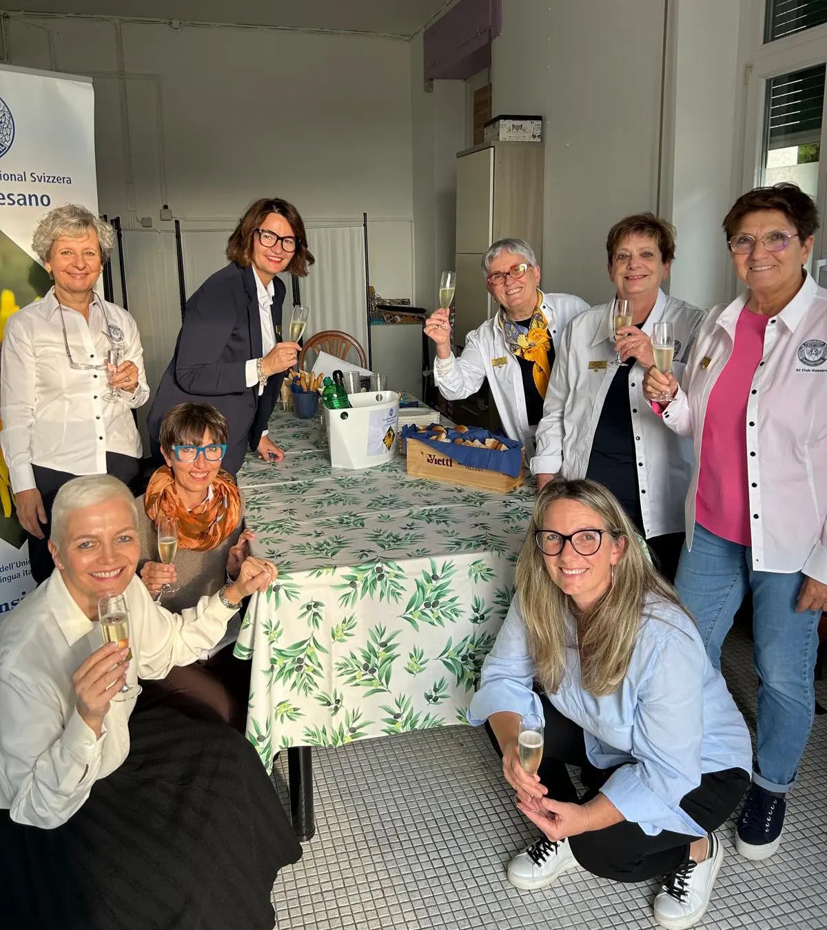 Un gruppo di otto donne sorridenti si trova attorno a un tavolo decorato con una tovaglia a foglie. Alcune di loro brindano con flute di champagne, mentre una ragazza con occhiali sorride al centro. Sul tavolo ci sono bottiglie di vino e un cesto. Una bandiera svizzera è visibile sullo sfondo.