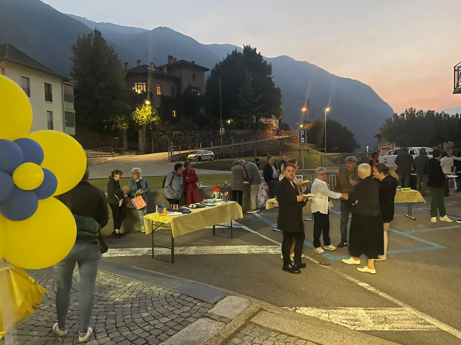 Un gruppo di persone partecipa a un evento all'aperto di sera, con tavoli allestiti e decorazioni gialle e blu. Sullo sfondo si vedono montagne e luci delle case.