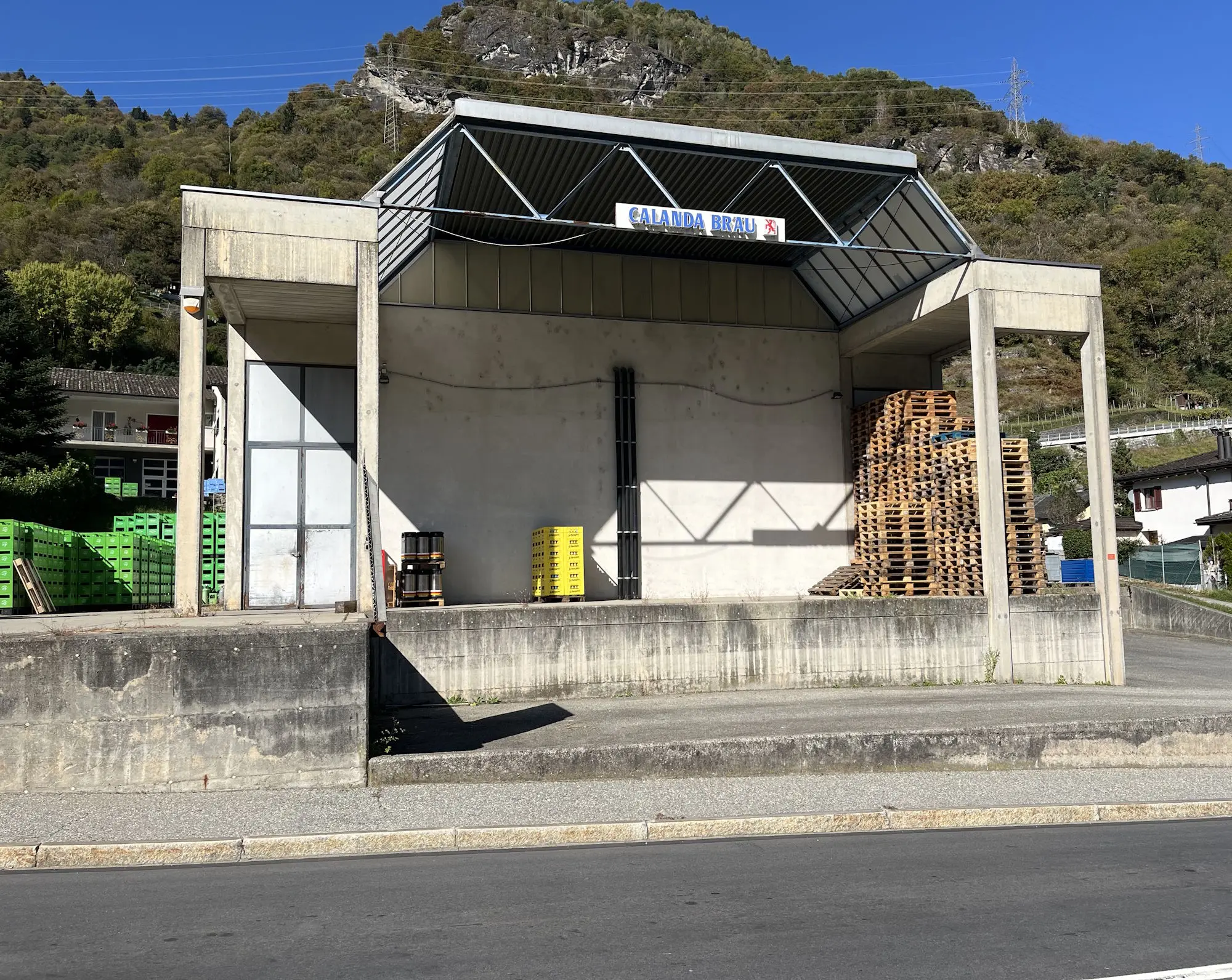 Un edificio vuoto con un tetto a spiovente, caratterizzato da una facciata di cemento e pareti chiare. Sul pavimento, ci sono pallet di legno impilati e contenitori di plastica colorati. Sullo sfondo, si vede una collina coperta di alberi e un cielo sereno.