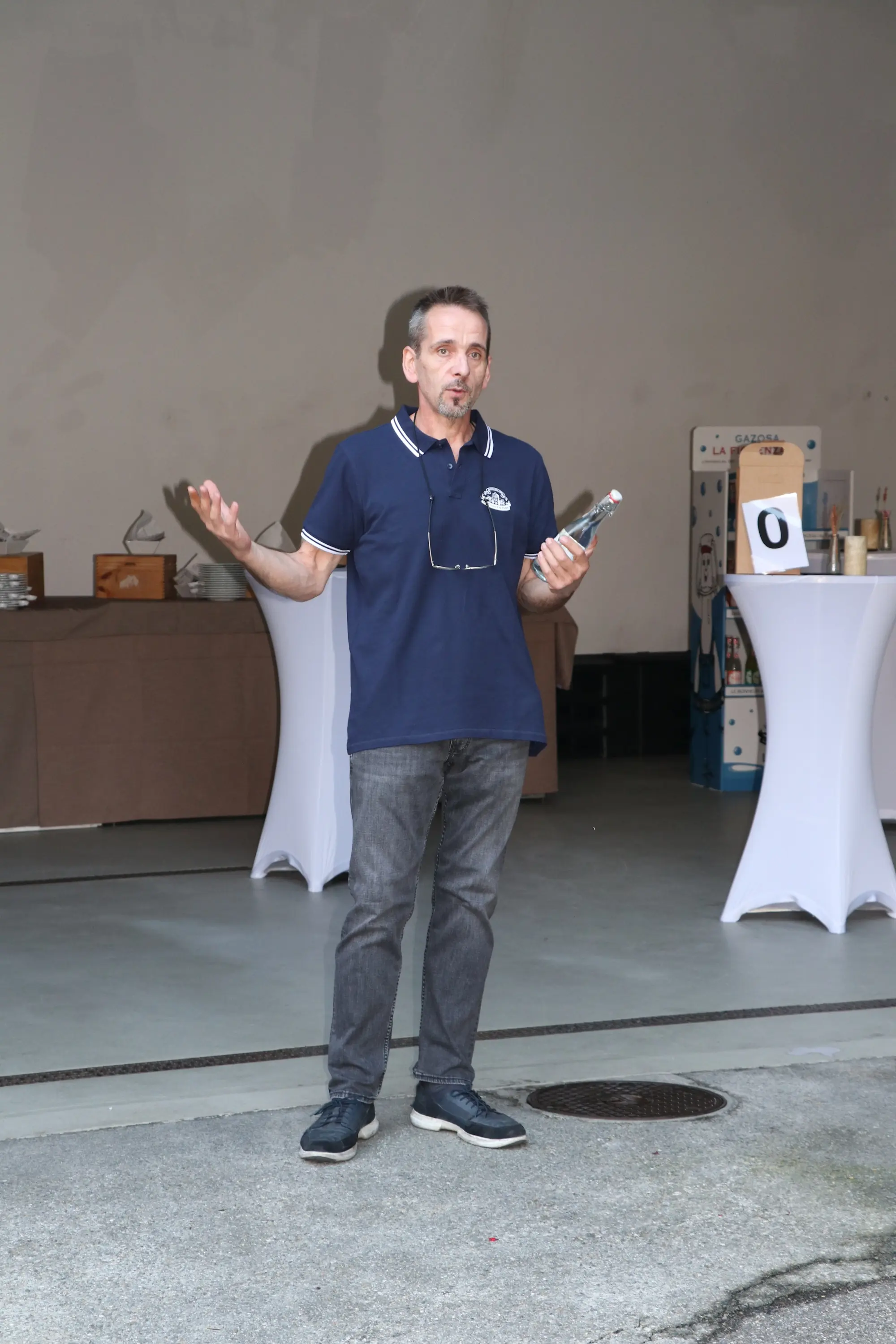 Un uomo in piedi, con una bottiglia in mano, sta parlando con gesti espressivi. Indossa una polo blu e pantaloni grigi, mentre sullo sfondo si vedono tavoli apparecchiati e una macchina per bibite.