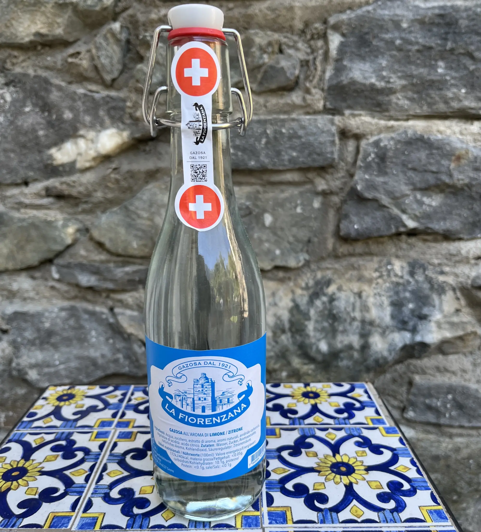 Bottiglia di acqua minerale Fiorenzana con etichetta blu e simbolo svizzero, appoggiata su una superficie rocciosa vicino a una piastrella decorata con motivi gialli e blu.