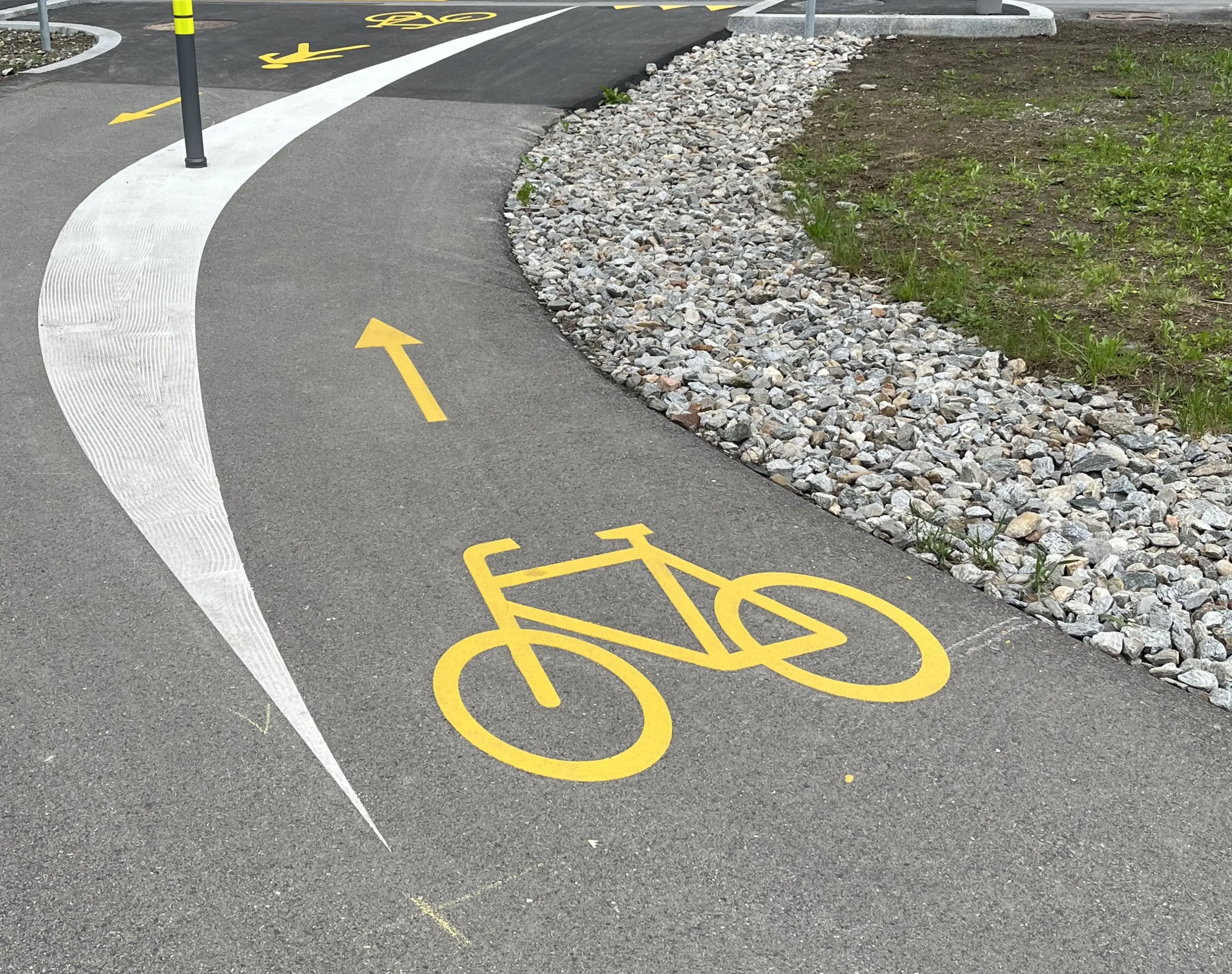 Un percorso ciclabile con segnali gialli sulla superficie in cemento, inclusa un'icona di bicicletta e una freccia che indica la direzione da seguire. Accanto, ci sono ciottoli decorativi e un'area verde.