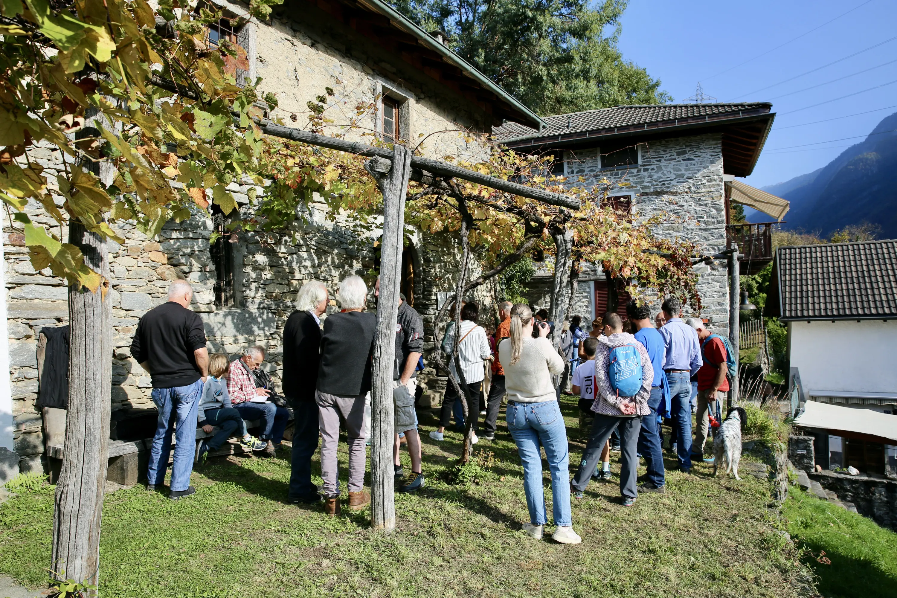 Un gruppo di persone si riunisce all'aperto vicino a case in pietra, circondato da viti e vegetazione. Alcuni partecipanti sono seduti, mentre altri chiacchierano in piedi. Si possono vedere montagne sullo sfondo.