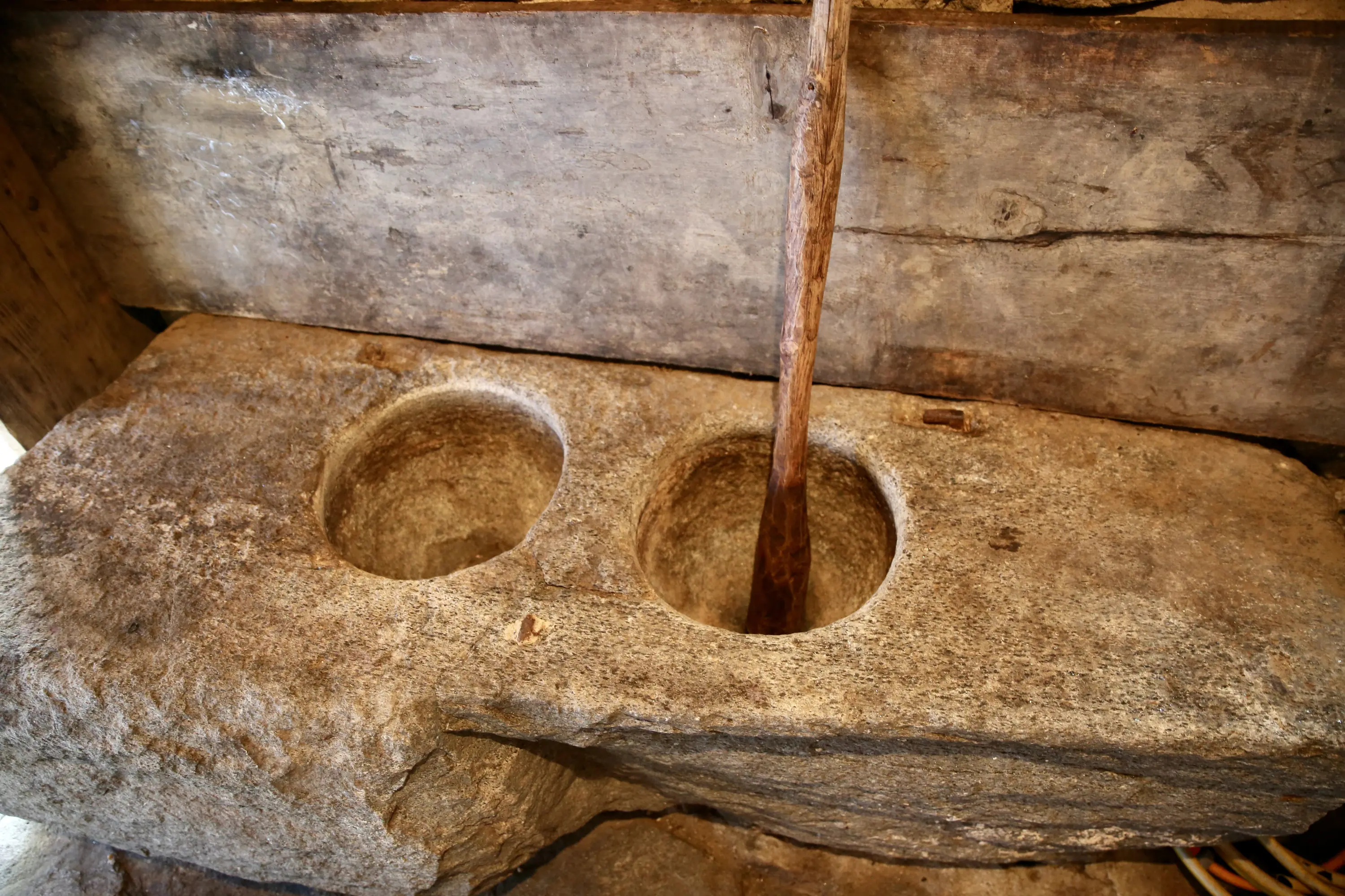 Due vasche di pietra, scolpite nella roccia, con un bastone di legno appoggiato sopra. La superficie è ruvida e mostra segni di usura, incastonata in un contesto di legno naturale.