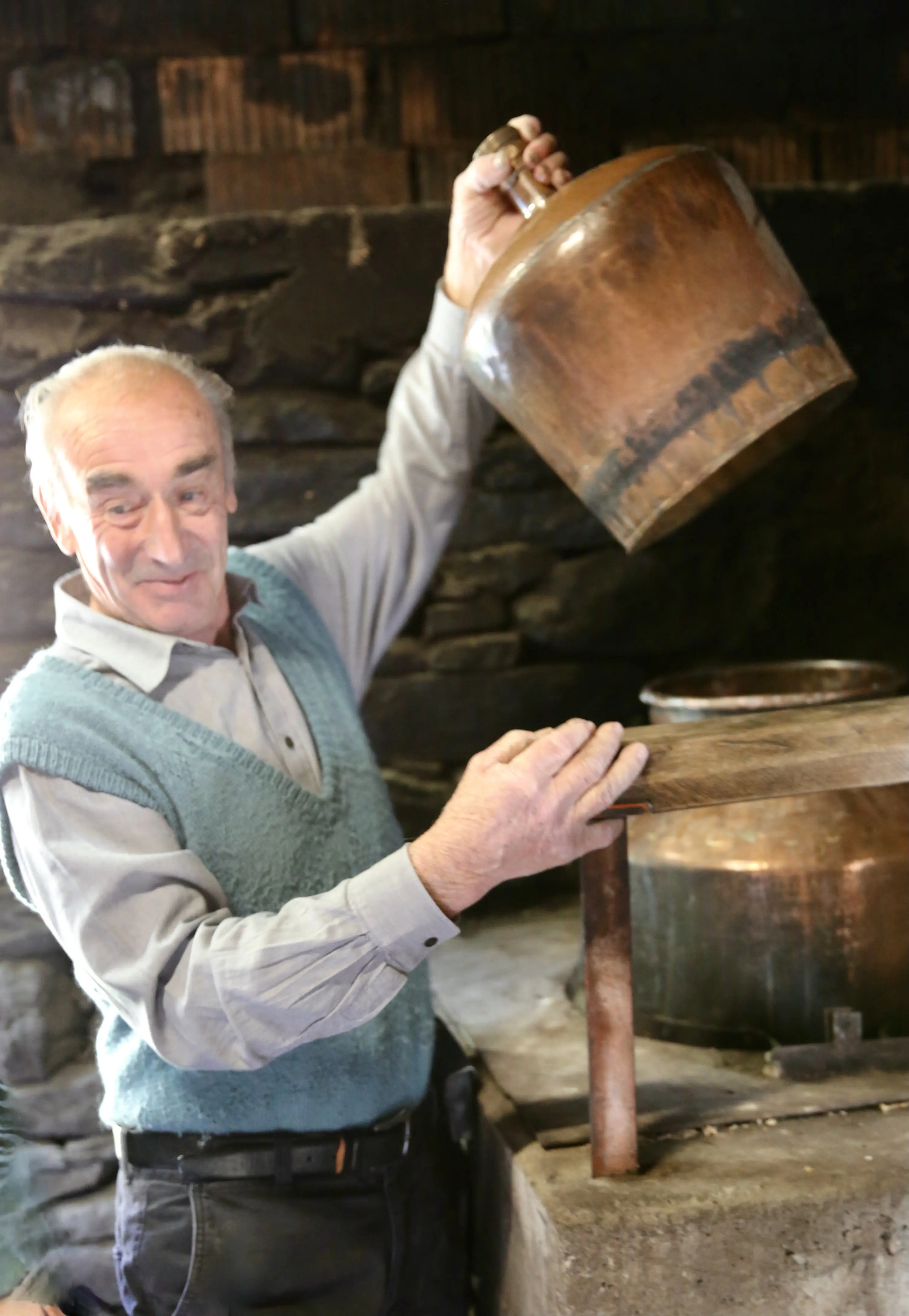 Uomo anziano con un maglione azzurro e camicia grigia, sorregge una pentola di rame sopra un tavolo di legno in un ambiente rustico. La scena trasmette un senso di tradizione e lavorazione artigianale.
