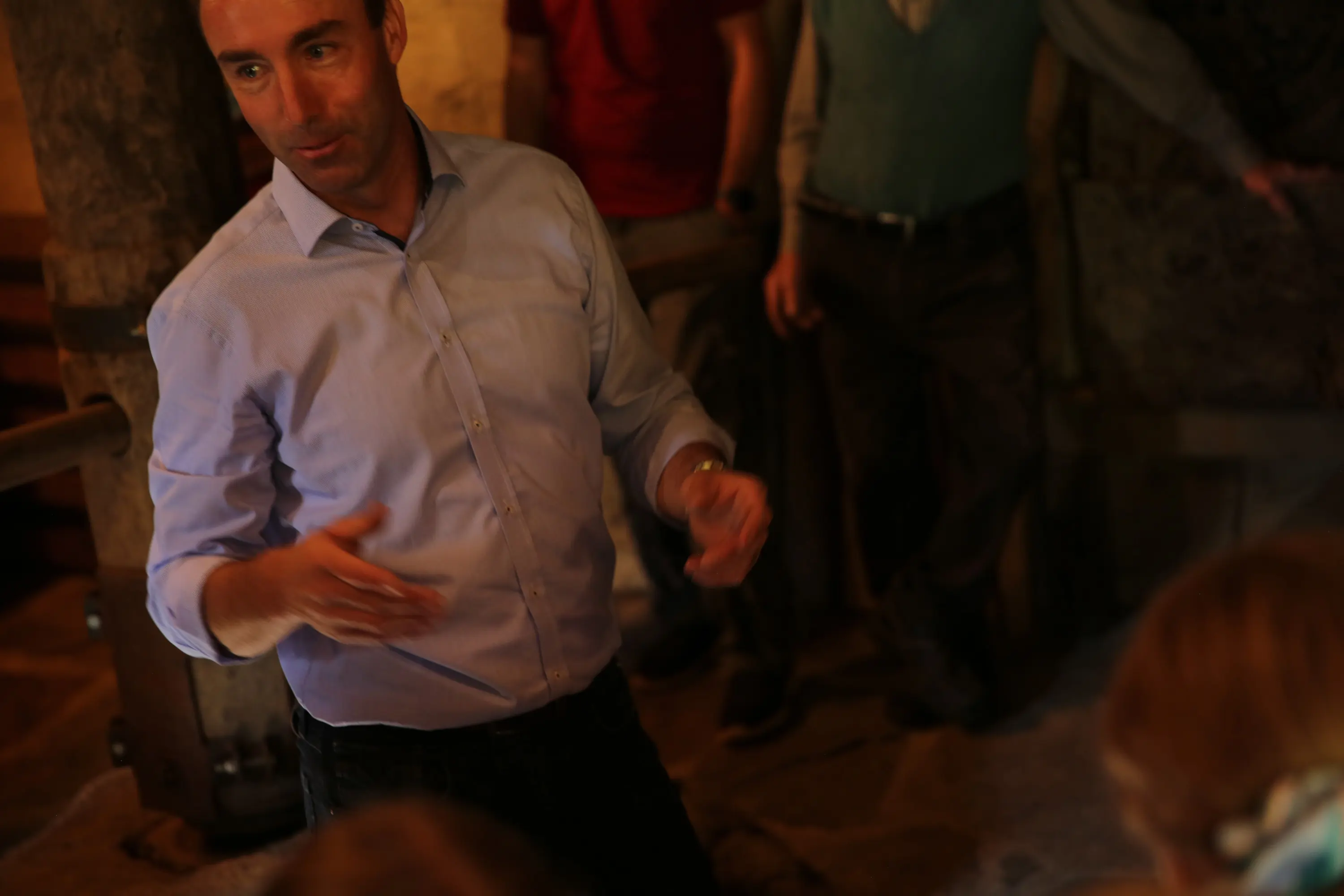 Un uomo con una camicia azzurra parla animatamente in un ambiente rustico, circondato da altre persone in secondo piano.