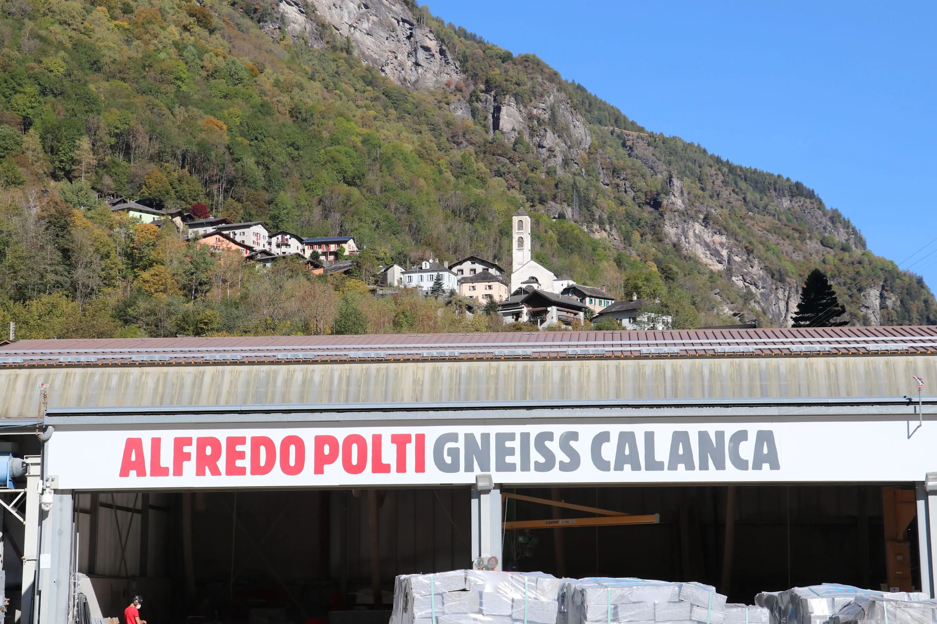 Un capannone con il nome "Alfredo Polti Gneiss Calanca" in evidenza, davanti a una vallata montuosa fitta di alberi. Sullo sfondo, si intravedono edifici e una chiesa, tipici di un paesaggio montano. Il cielo è sereno e blu.